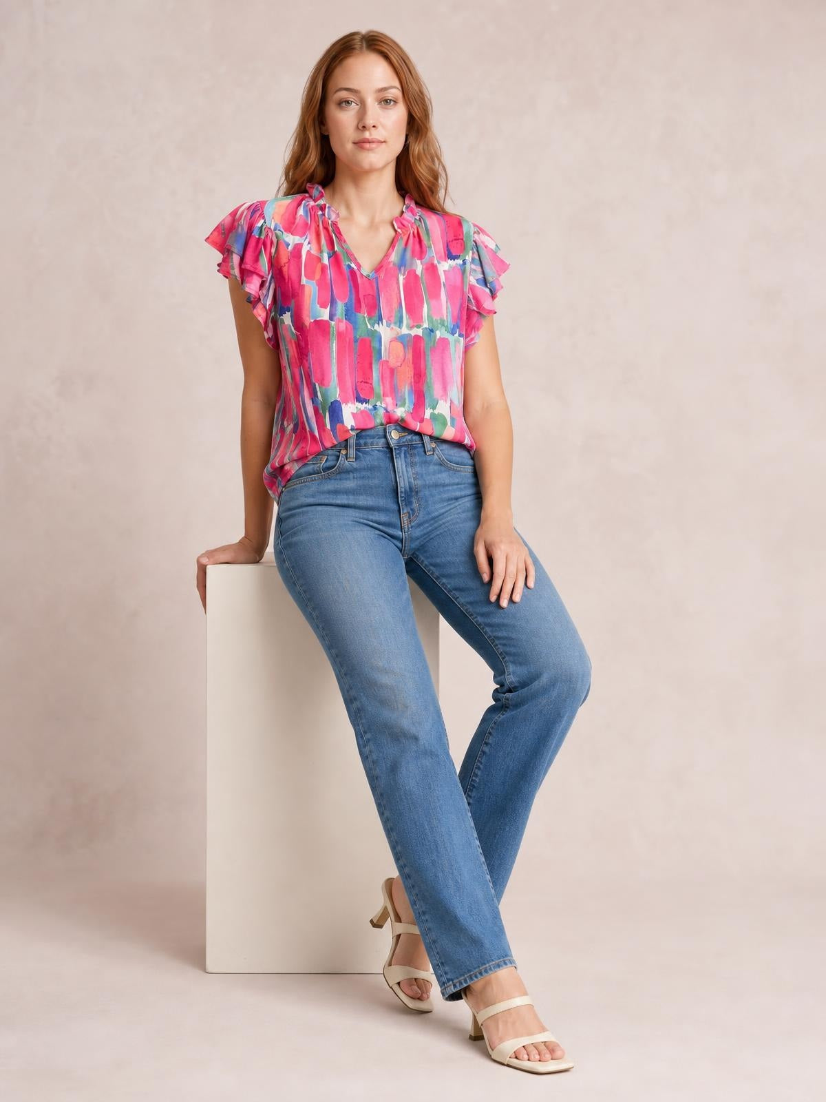A model sits in a colorful printed blouse with ruffled cap sleeves, a notched V-neckline, and a relaxed silhouette, paired with blue jeans.