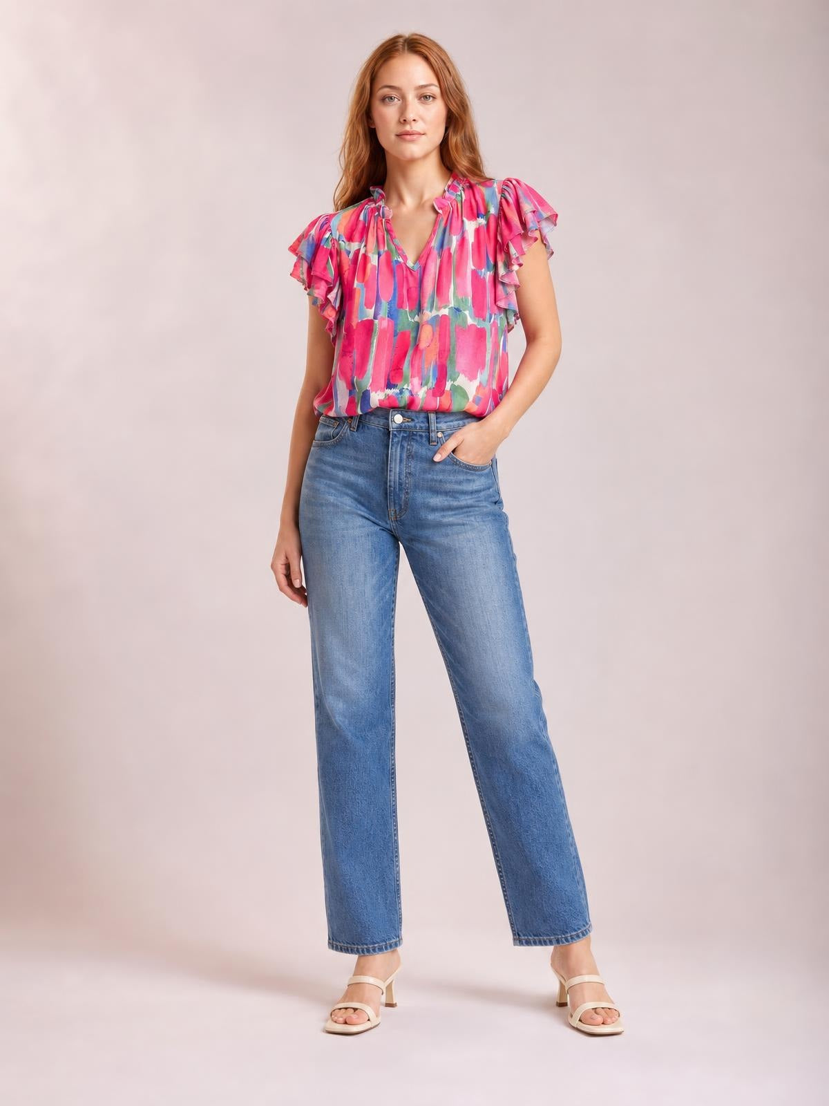 Model wearing a colorful printed blouse with ruffled cap sleeves, a notched V-neck, and a relaxed straight silhouette, posed with one hand in her jeans pocket.