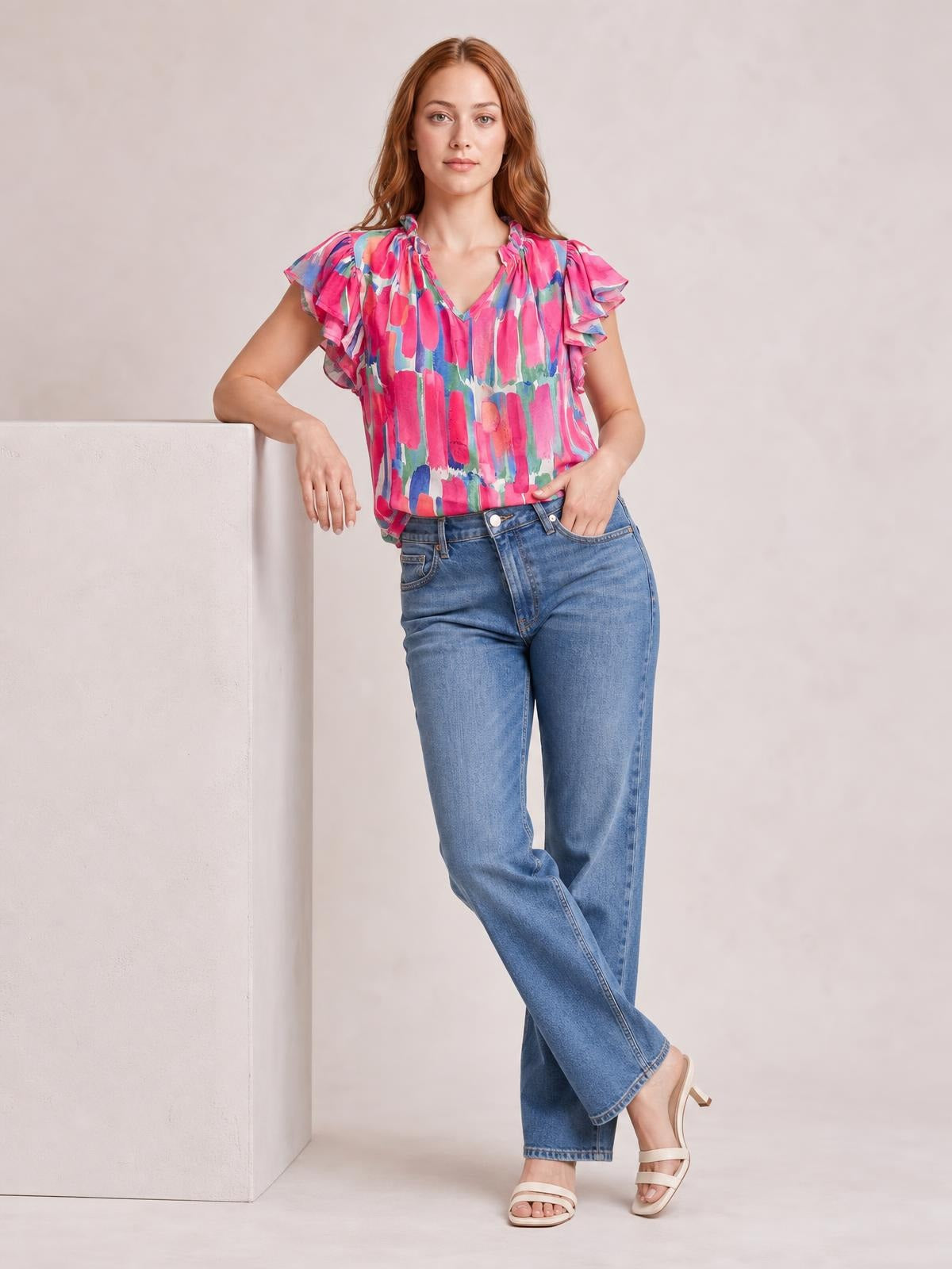 A model wears a colorful printed blouse with ruffled cap sleeves and a notched neckline, styled in a relaxed silhouette with one hand in her pocket.