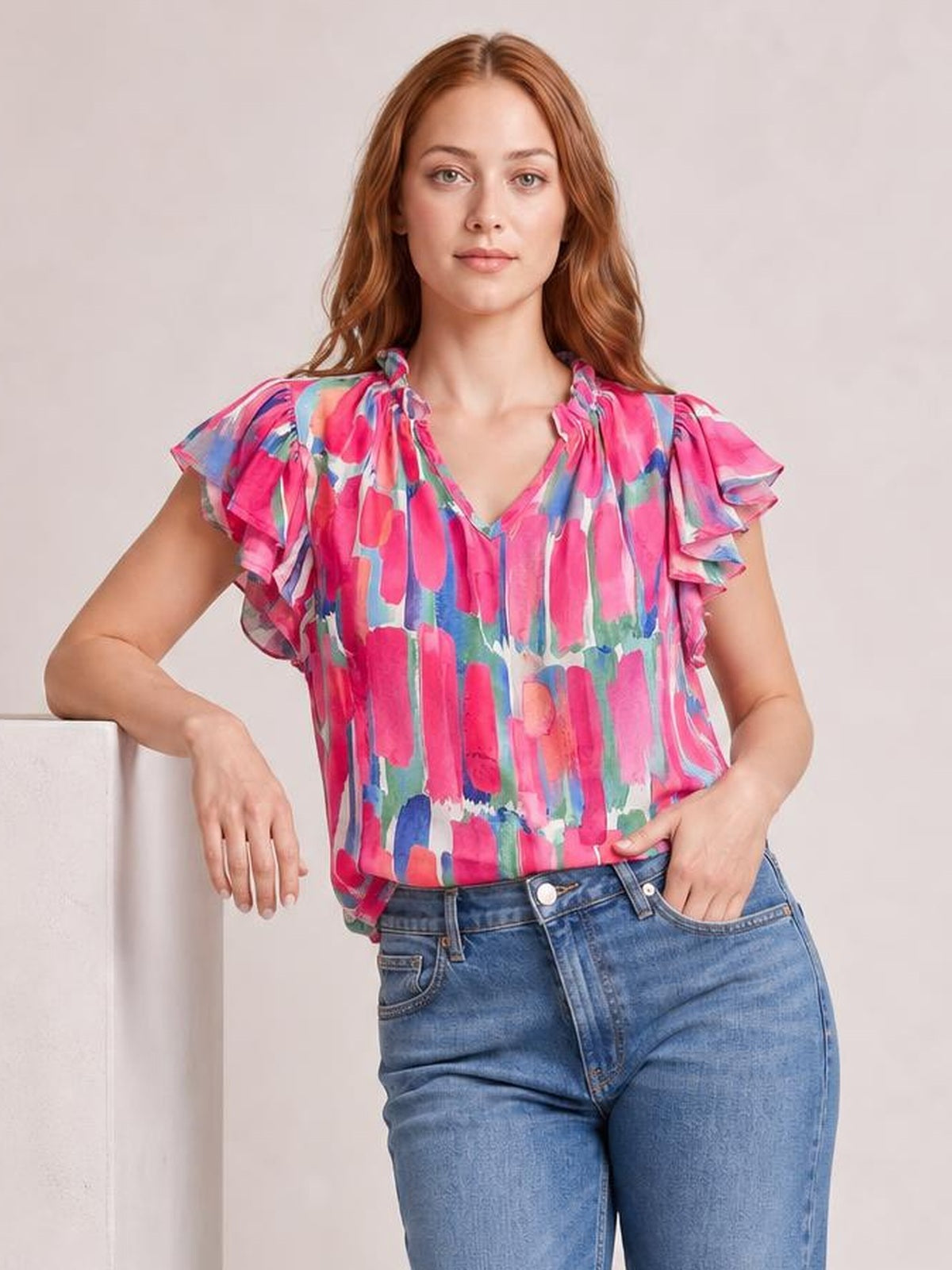 A model poses with one hand in her jeans pocket, wearing a colorful printed blouse with a notched neckline, ruffled cap sleeves, and a relaxed silhouette.
