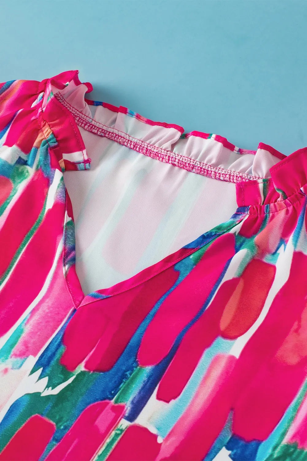A vibrant printed blouse in hot pink, blue, green, and white features a ruffled notched neckline, short cap sleeves, and a relaxed silhouette.
