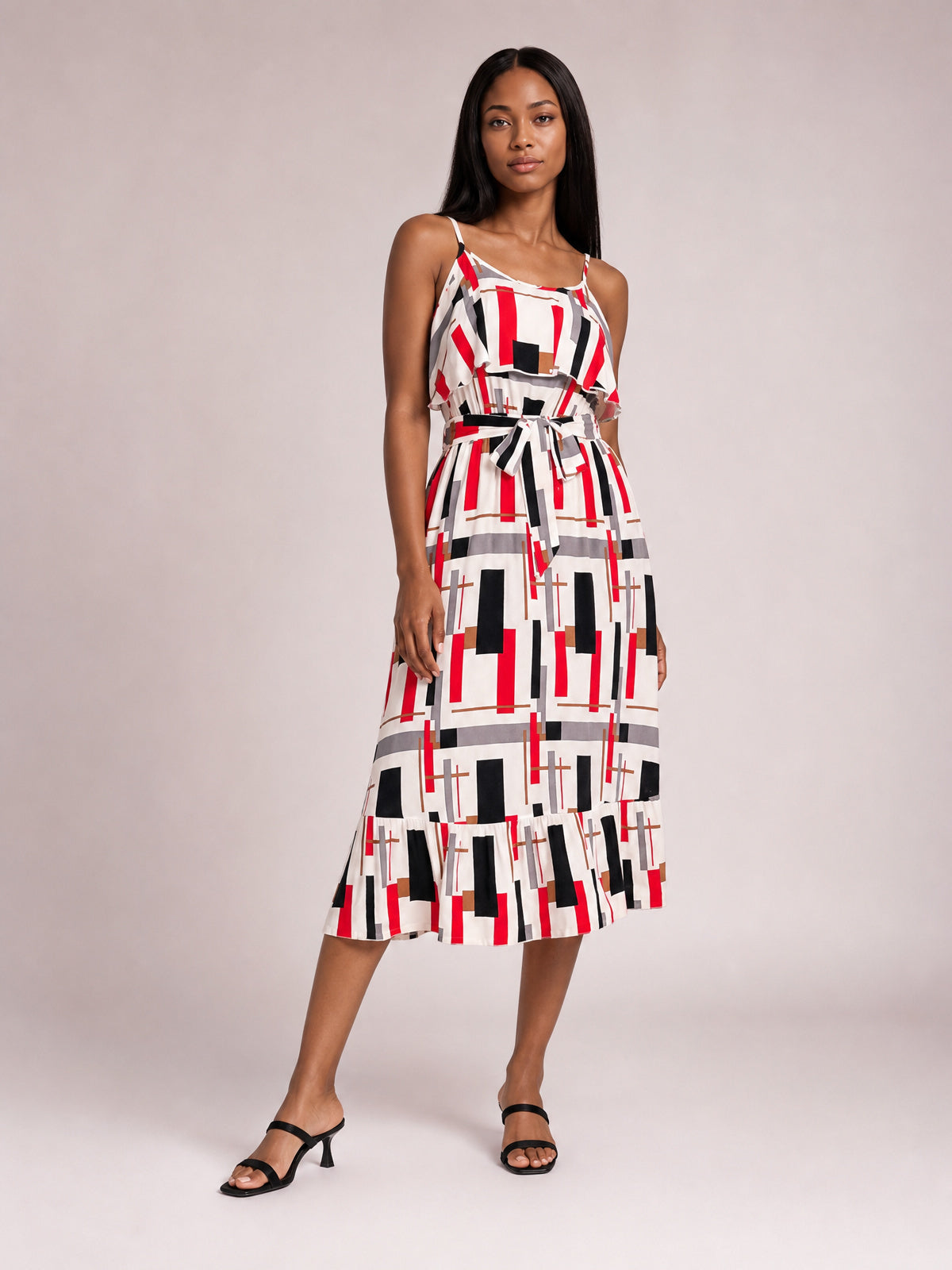 Model poses in a geometric multi-color printed midi dress with slender straps, ruffled bodice overlay, self-tie waist, and flared hem.