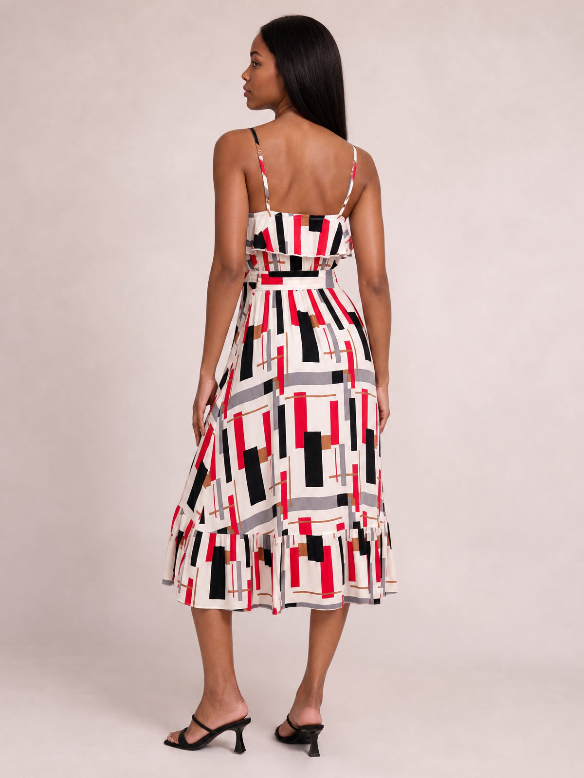 Model in a back view wears a multicolor geometric printed midi dress with slender straps, ruffled bodice overlay, tied waist, and gently flared tiered skirt.