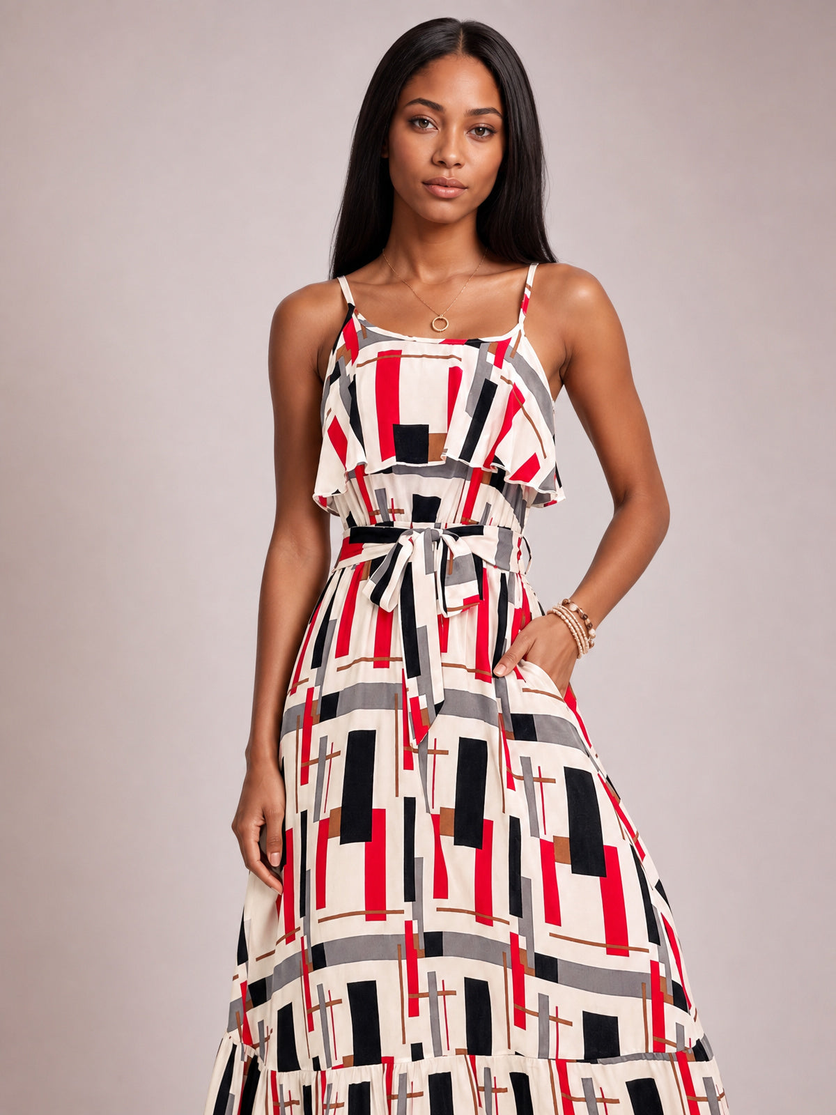 A model poses in a multi-color geometric printed midi dress with slender shoulder straps, a ruffled bodice overlay, defined tie waist, and gently flared silhouette.
