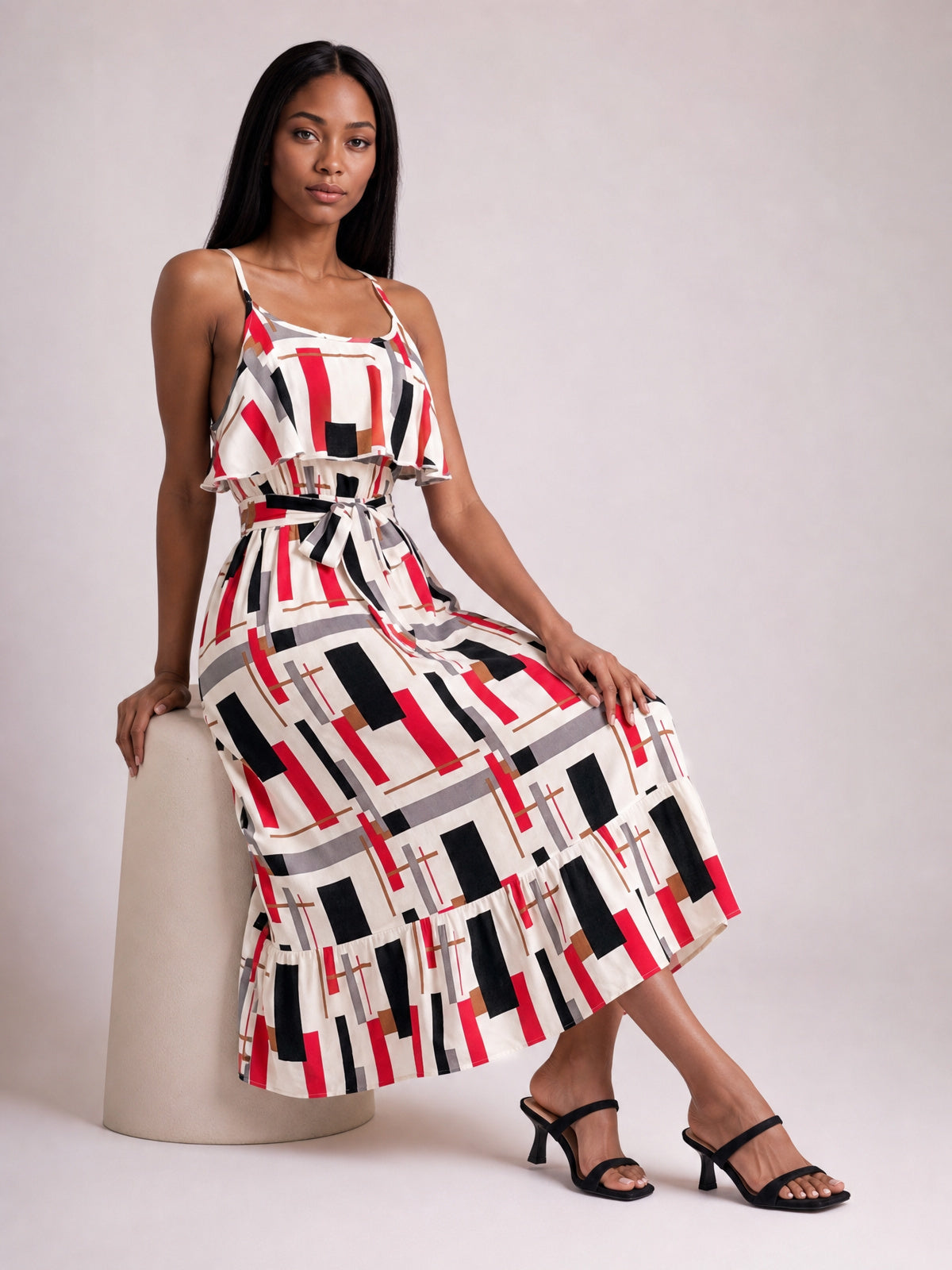 A model leans against a seat wearing a multicolor geometric printed midi dress with slender straps, a ruffled bodice overlay, tie waist, and flared skirt.