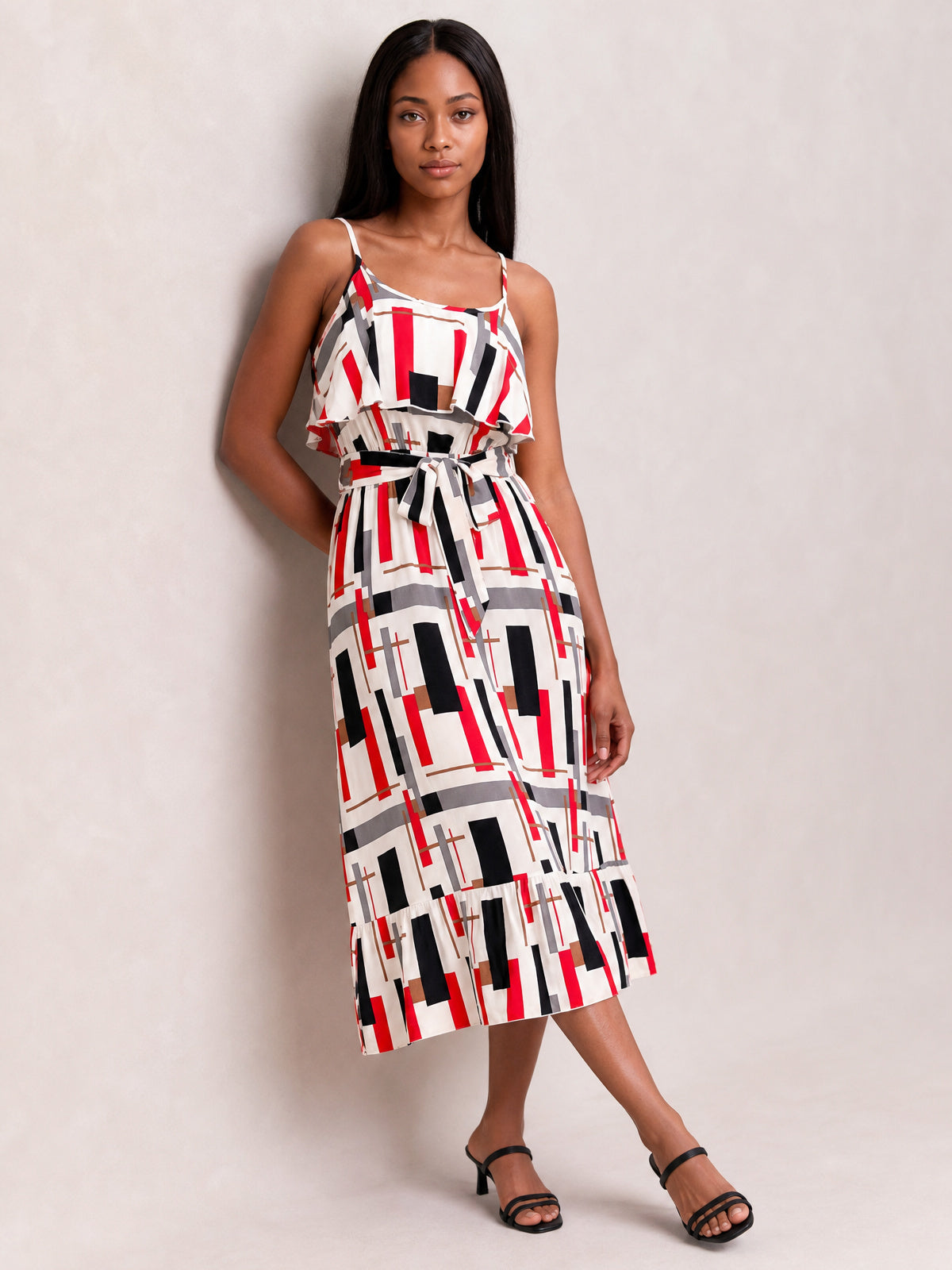 Model poses in a geometric printed midi dress with red, black, gray, and white tones, slender straps, ruffled bodice overlay, tie waist, and flared hem.