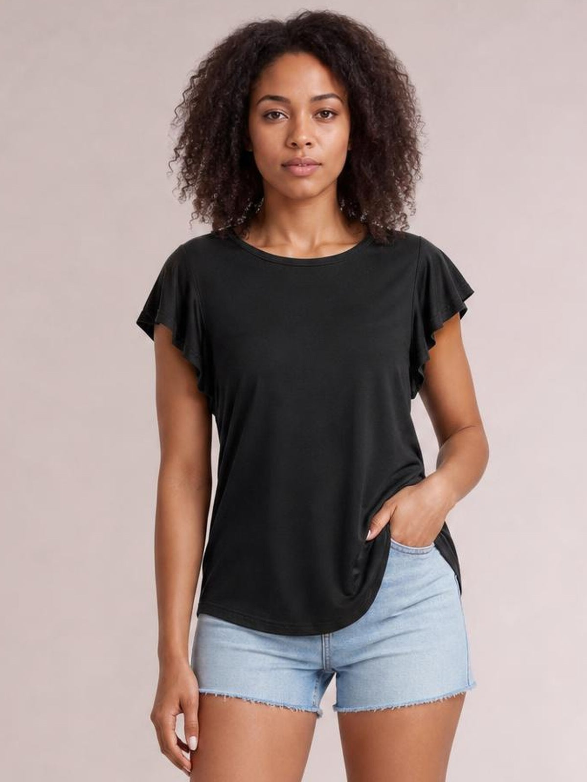 Model wearing a black ruffled round neck cap sleeve blouse with a relaxed straight silhouette, standing with one hand in her shorts pocket.