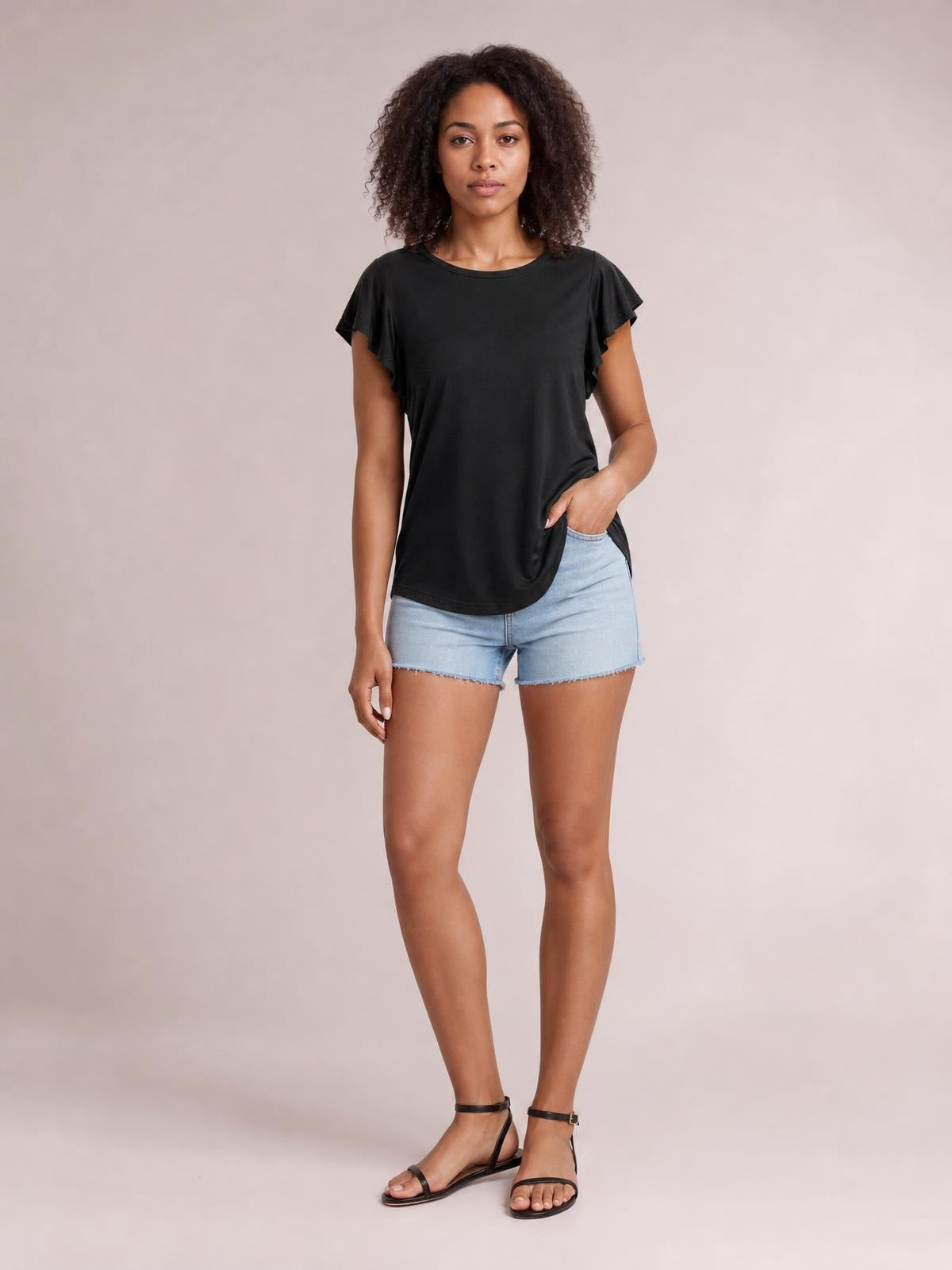 A model wears a black ruffled round neck cap sleeve blouse with a relaxed silhouette, standing with one hand in her shorts pocket.