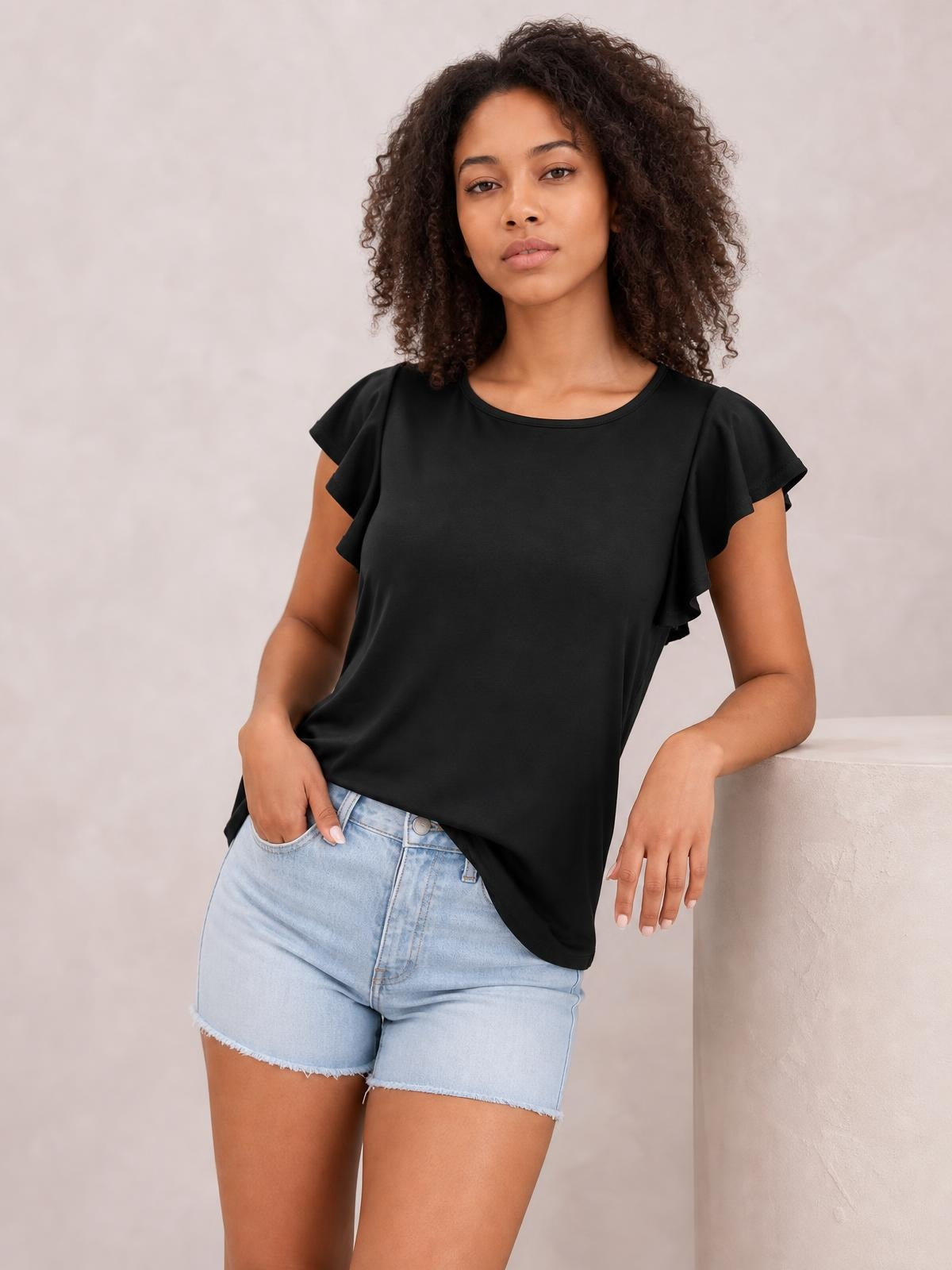 Model wearing a black ruffled round neck cap sleeve blouse with a relaxed silhouette, standing with one hand in her shorts pocket.