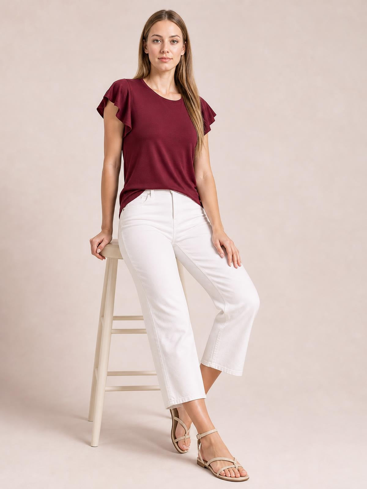 A model wears a burgundy ruffled round neck cap sleeve blouse with a relaxed silhouette, leaning against a stool in white cropped pants.