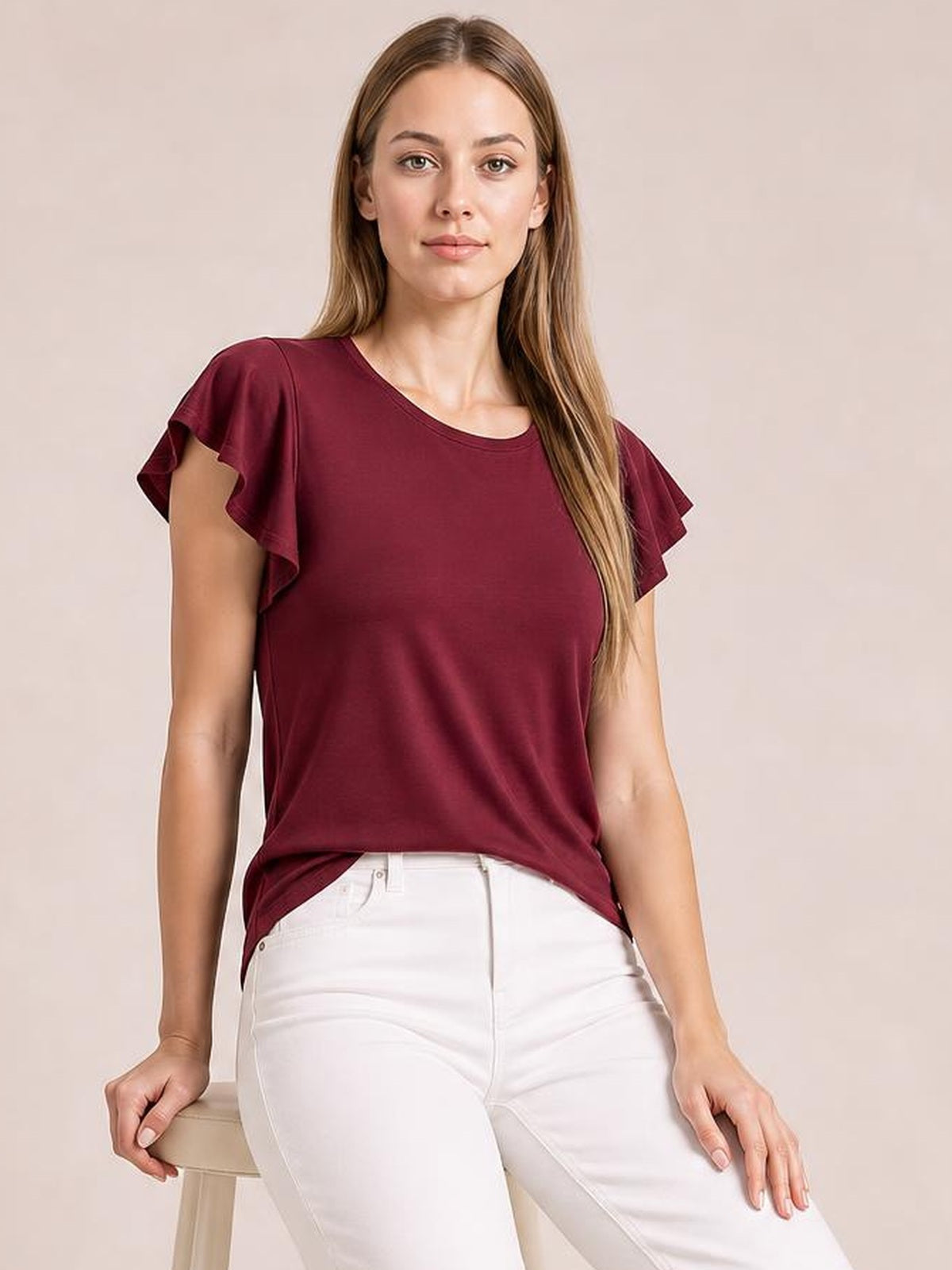 Model wearing a burgundy ruffled round-neck blouse with layered cap sleeves and a relaxed silhouette, leaning against a stool.