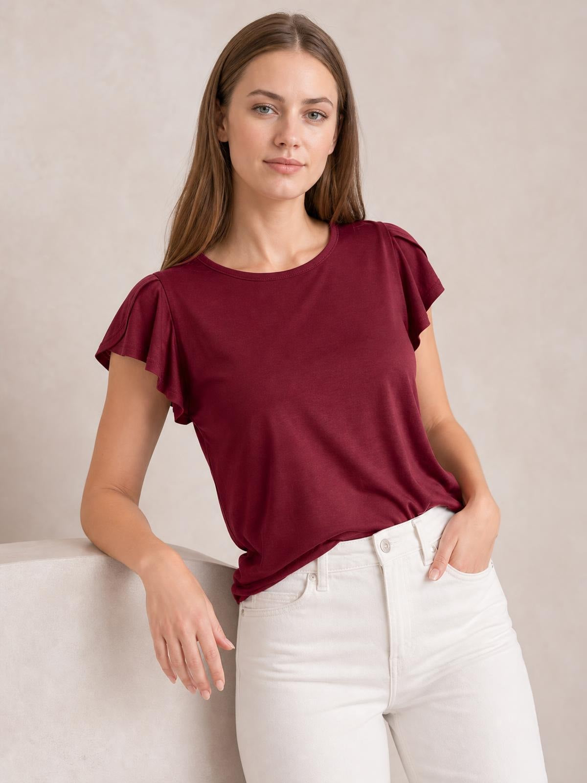 A model wears a burgundy ruffled round-neck blouse with fluttering cap sleeves, a relaxed silhouette, and a softly draped hem over white jeans.