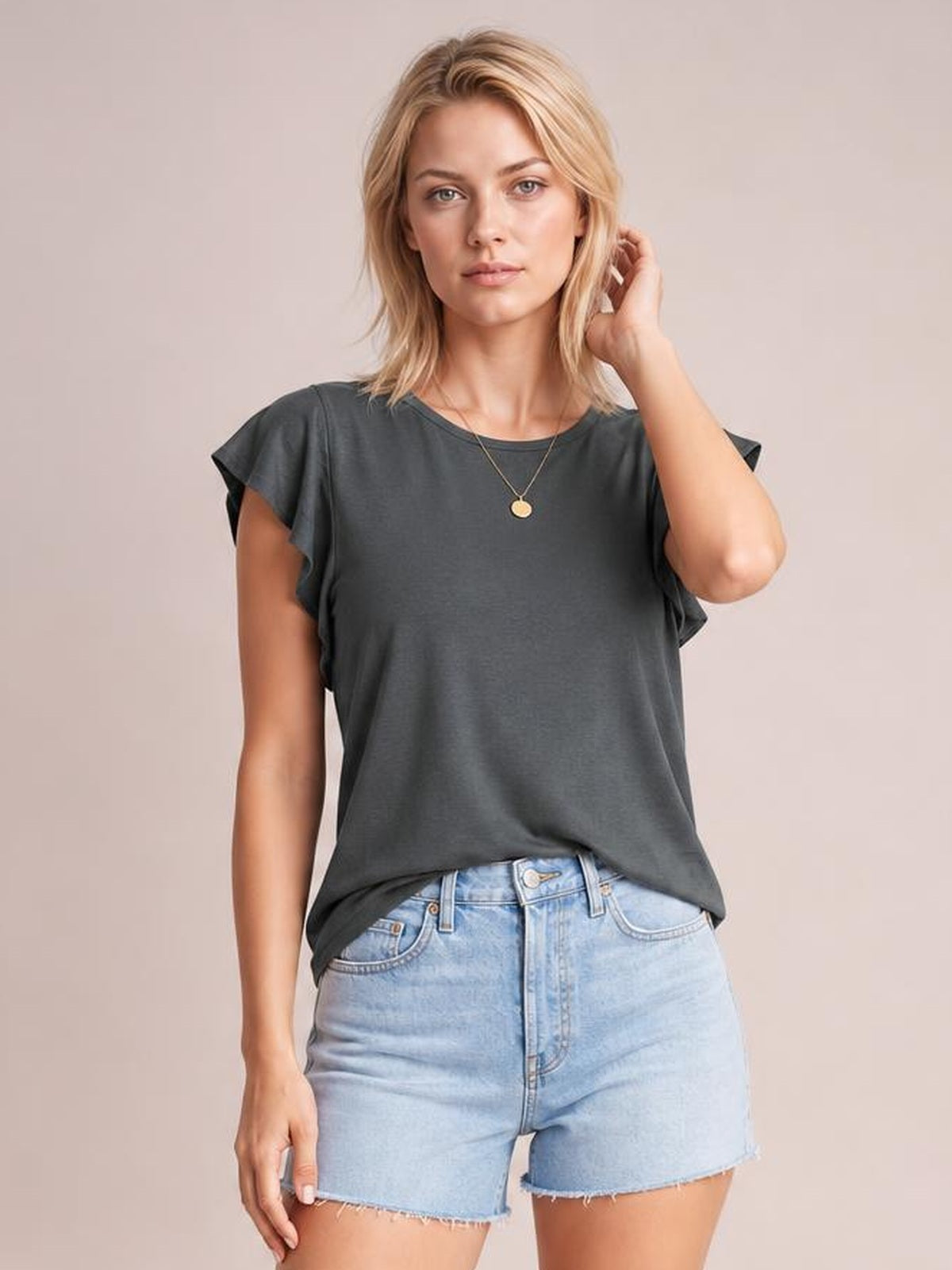 Model wearing a dark gray ruffled round-neck cap-sleeve blouse with a relaxed straight silhouette, standing front-facing with one hand touching her hair.