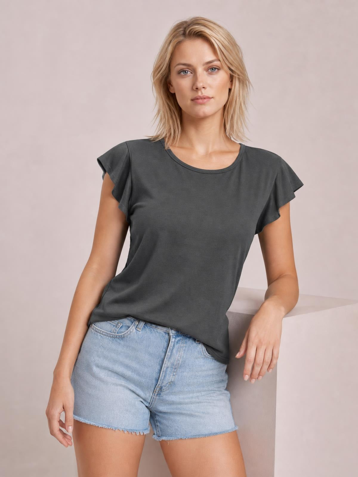A model leans against a ledge wearing a charcoal ruffled round-neck cap-sleeve blouse with a relaxed, slightly loose silhouette and soft drape.