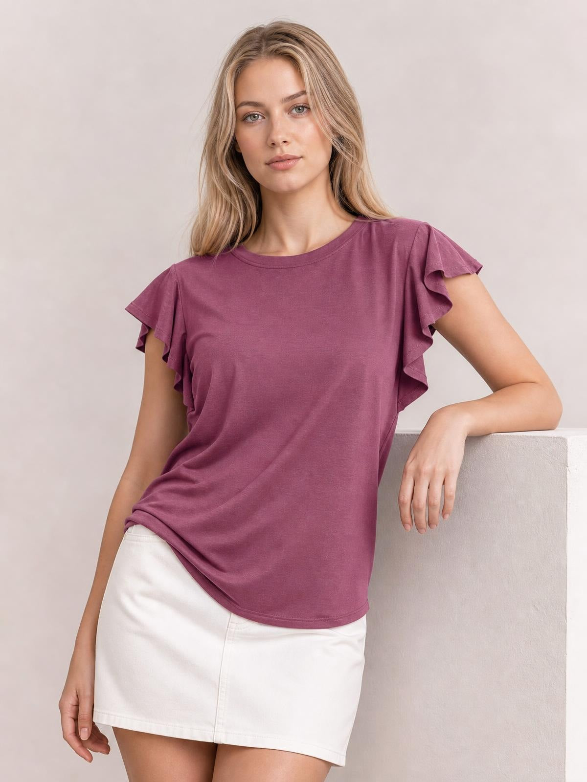 Model wears a mauve ruffled round neck cap sleeve blouse with a relaxed straight silhouette and curved hem, leaning casually with one arm supported.