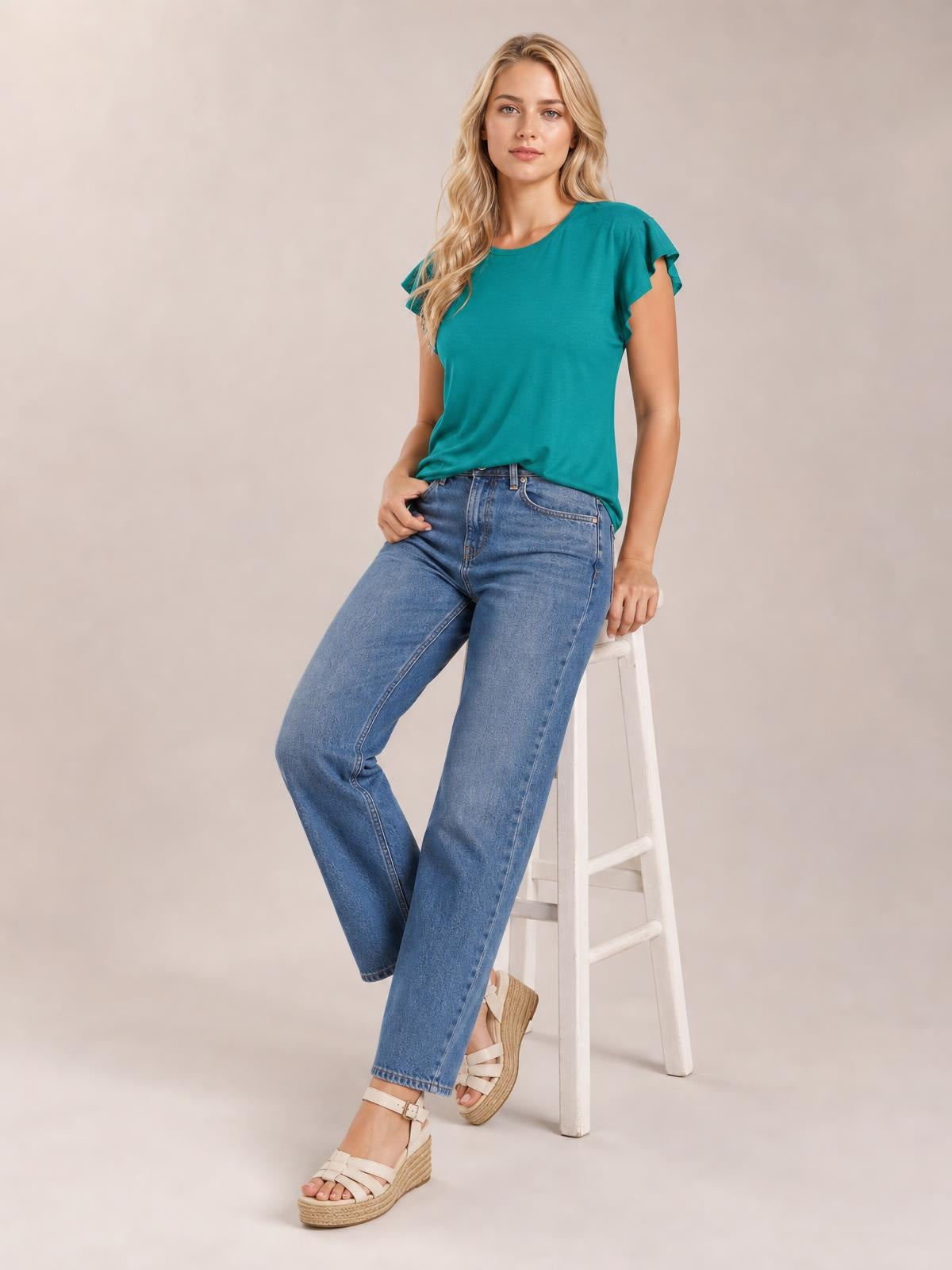 A model wears a teal ruffled round neck cap sleeve blouse with a relaxed silhouette, leaning against a white ladder in jeans and wedge sandals.