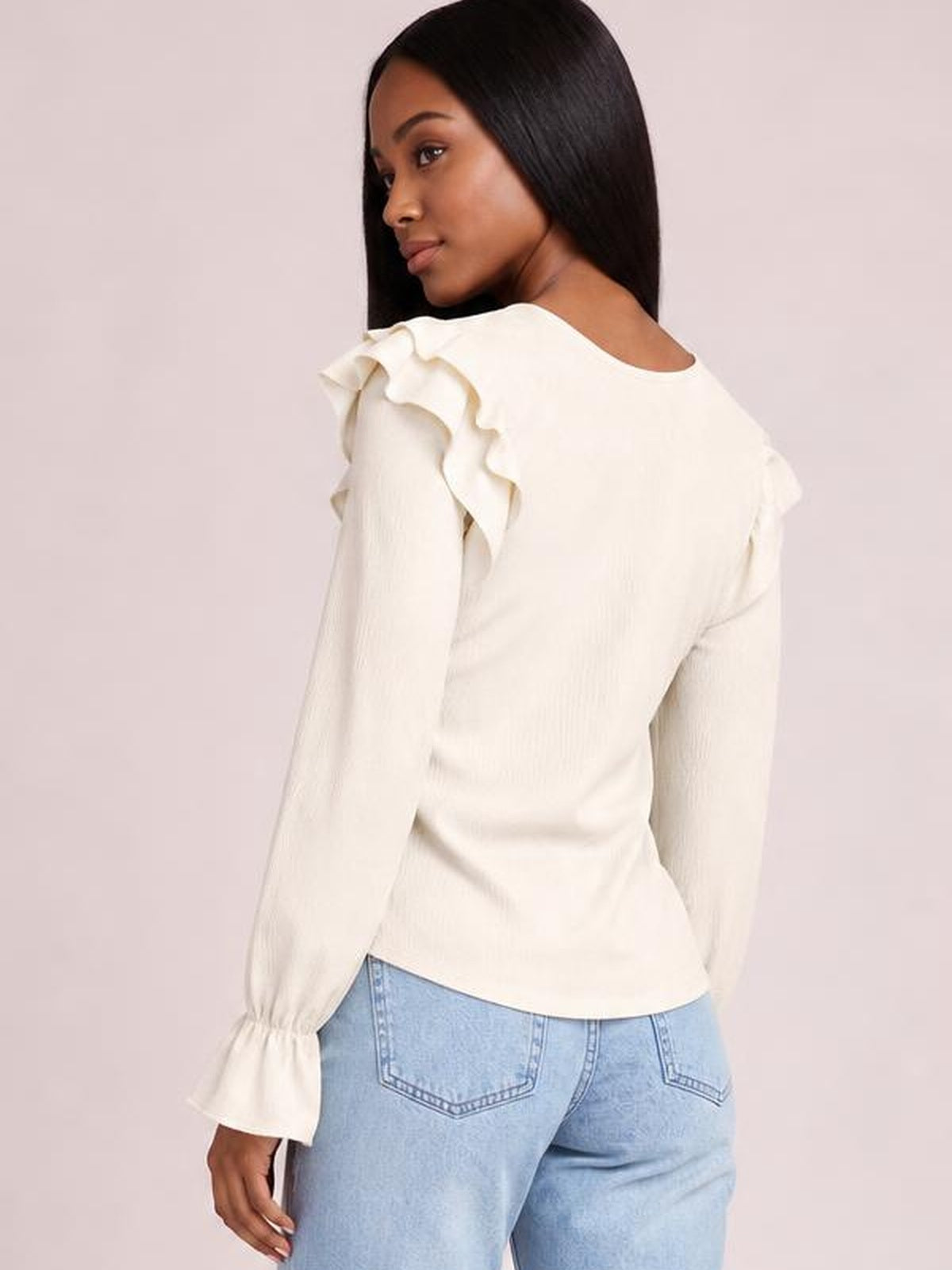 Model turned to show the back of a cream round-neck top with ruffled shoulder details, flounce sleeves, fitted silhouette, and gathered cuffs.