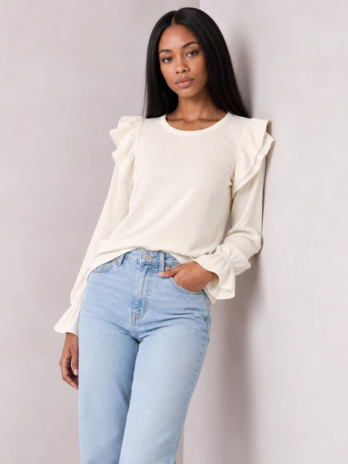 Model wearing an ivory round neck top with ruffled flounce sleeves, long cuffs, and a relaxed silhouette, posing with one hand in her jeans pocket.