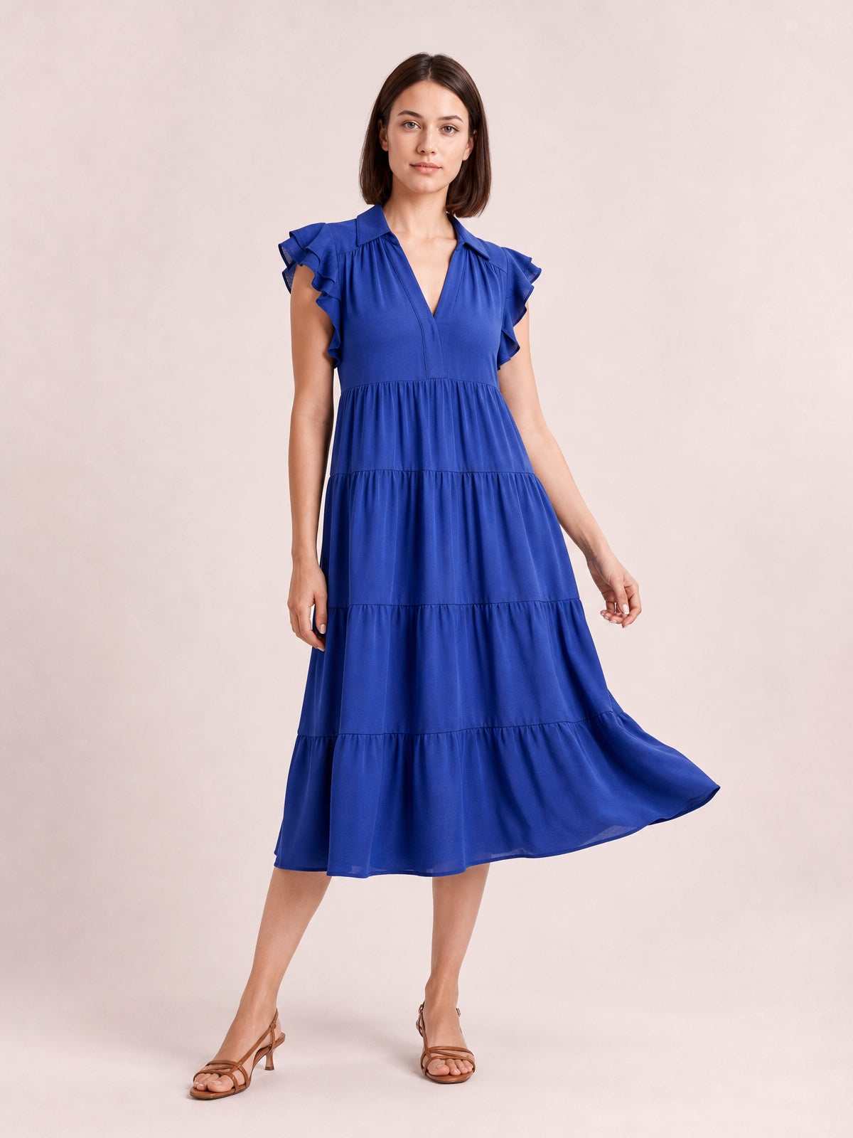Model poses in a rich blue ruffled short-sleeve dress with a collared V-neck, gathered bodice, tiered midi skirt, and flowing A-line silhouette.