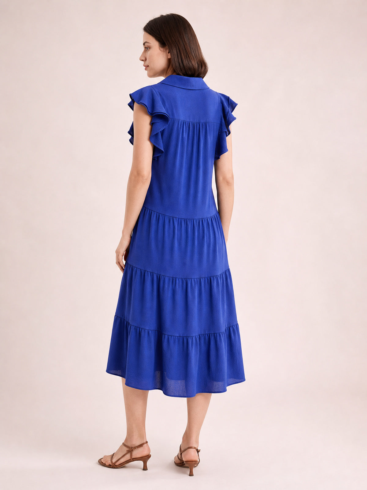 A model stands turned away in a rich blue collared midi dress with ruffled short sleeves, gathered seams, and a flowing tiered silhouette.