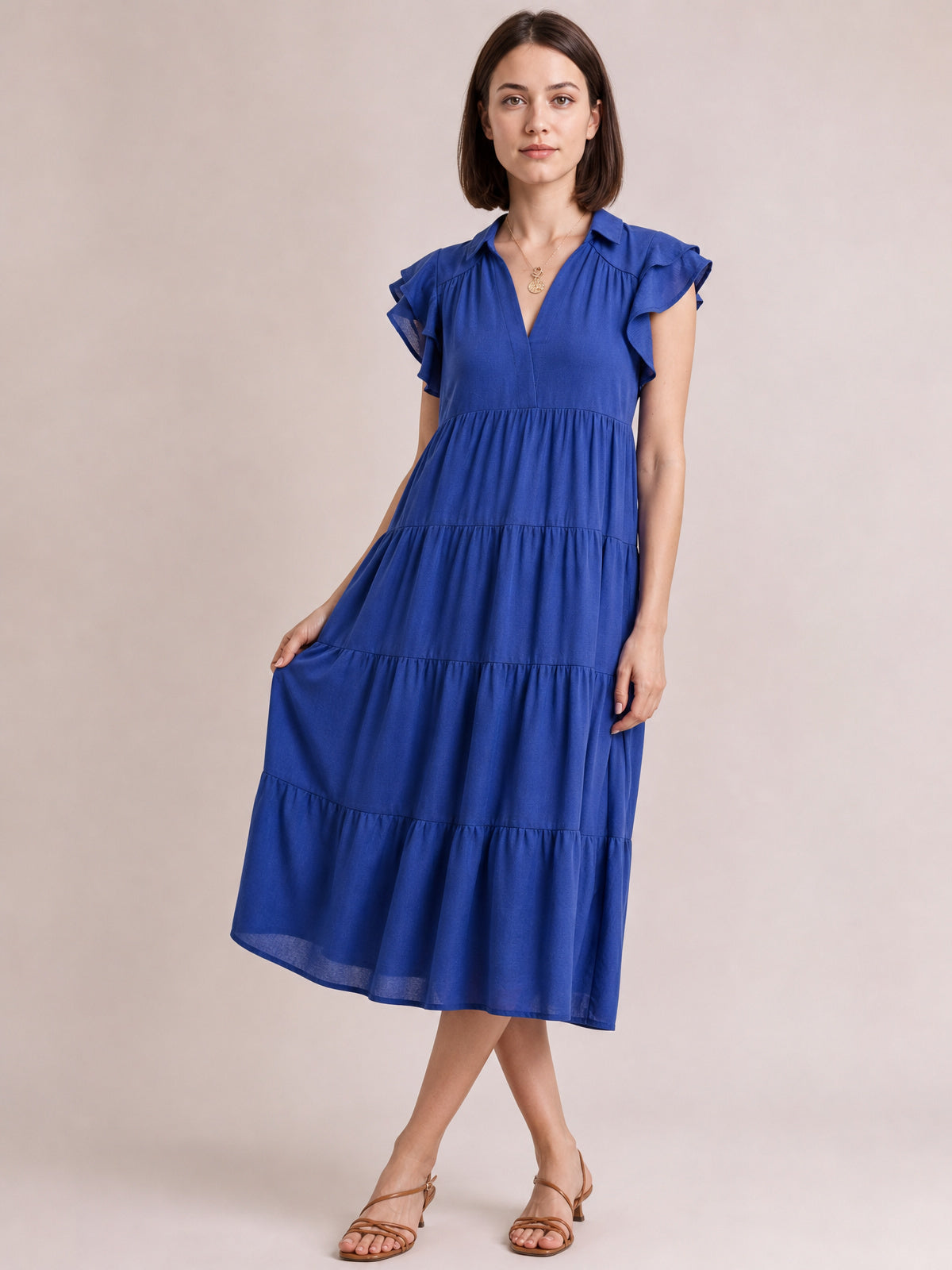Model poses in a light indigo tiered midi dress with a collared V-neck, ruffled short sleeves, gathered seams, and a flowing silhouette.