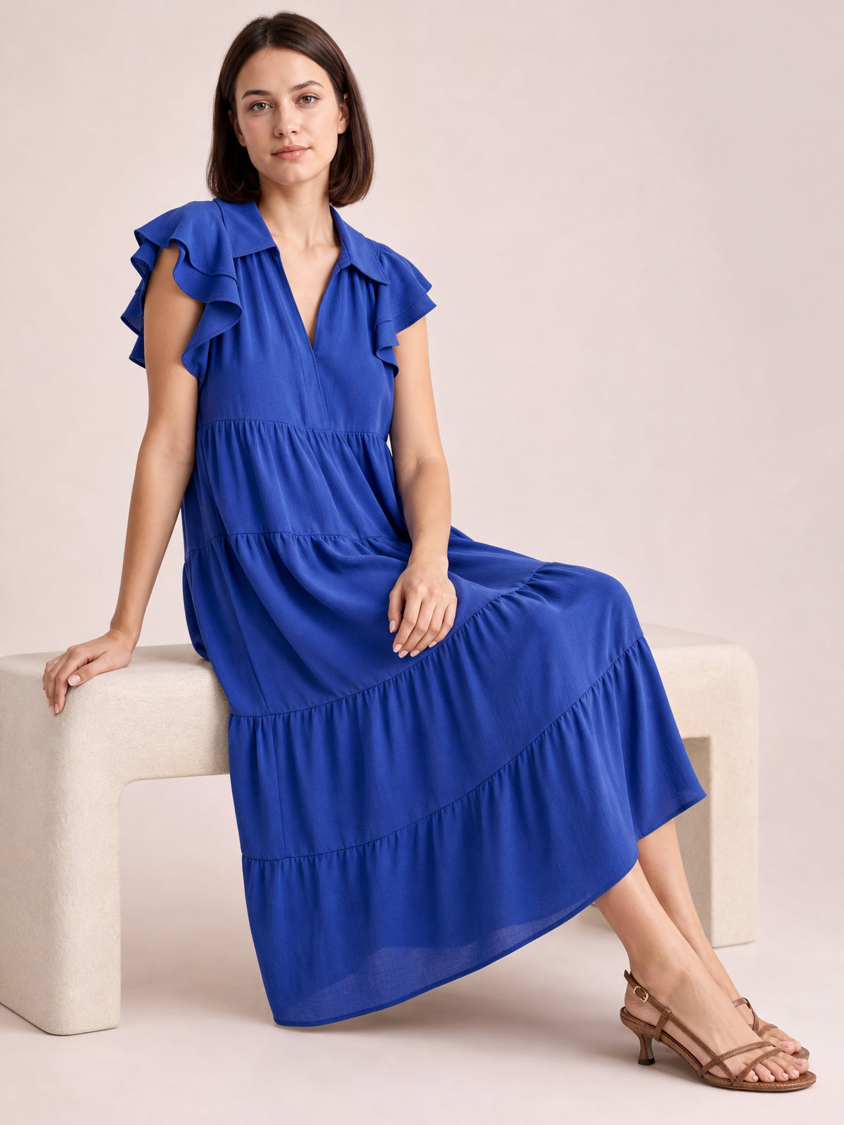 Model seated in a vivid blue midi dress with a collared V-neck, ruffled short sleeves, gathered seams, and a flowing tiered skirt.