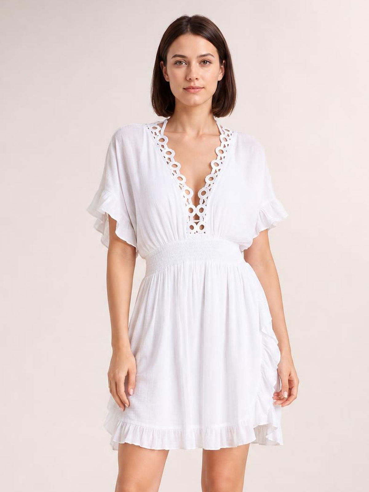 A model stands facing forward in a white V-neck cover-up with a smocked waist, ruffled sleeves and hem, and a decorative cutout neckline.