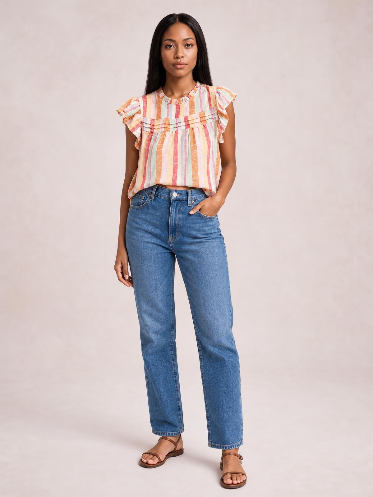 A model stands with one hand in her jeans pocket, wearing a loose striped blouse with a round neck, ruffled cap sleeves, and layered chest detailing.