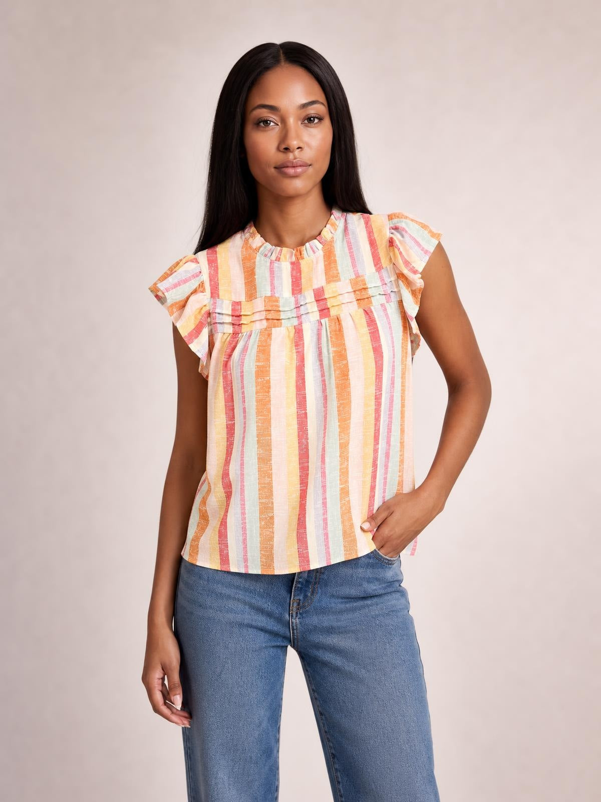 Model wearing a multicolor striped round-neck blouse with ruffled cap sleeves, layered ruffle detailing, and a relaxed straight silhouette, posing with one hand in her jeans pocket.