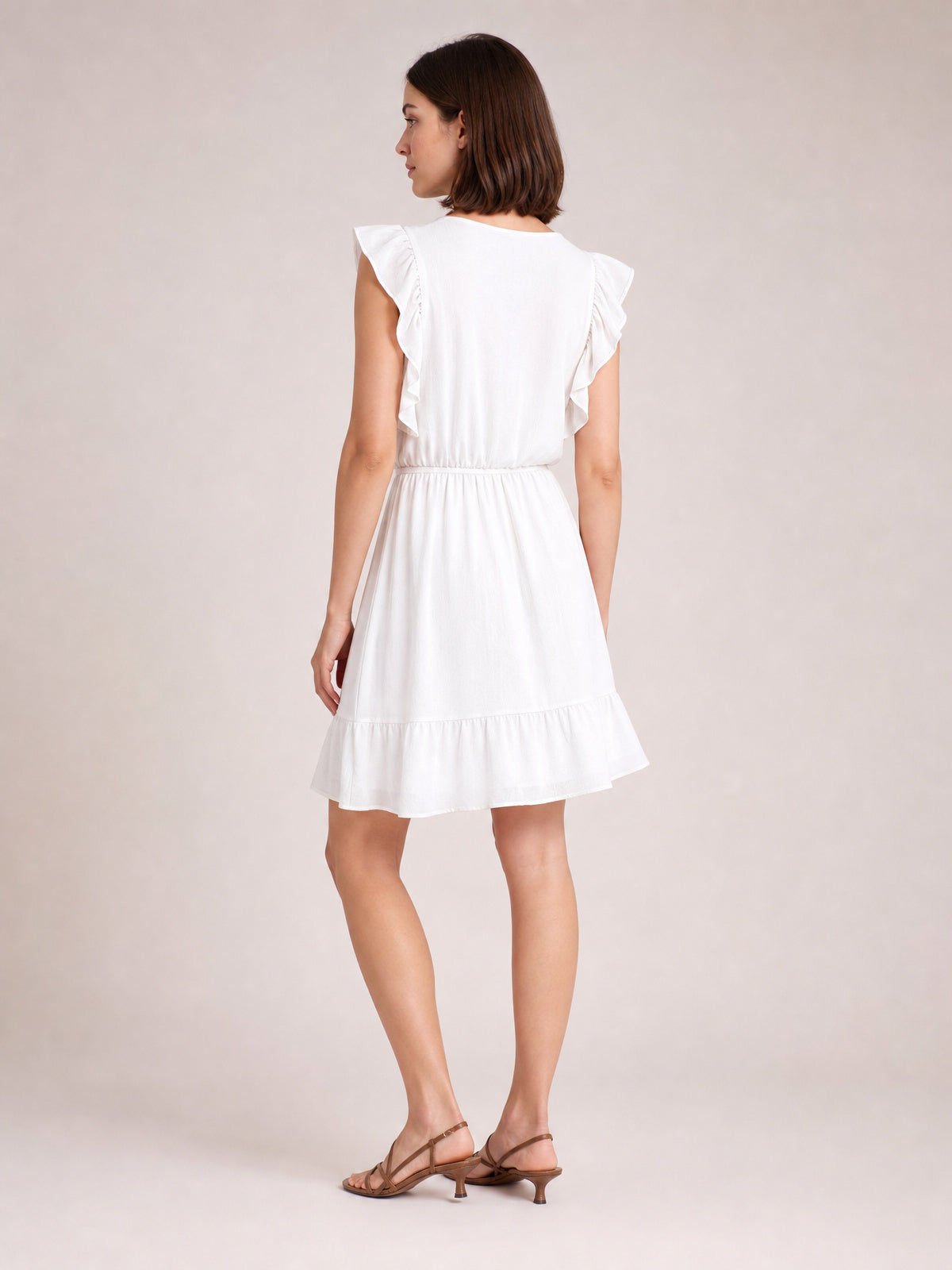 Model turned away in a white ruffled cap sleeve dress with a defined waist, softly gathered skirt, and tiered ruffle hem.