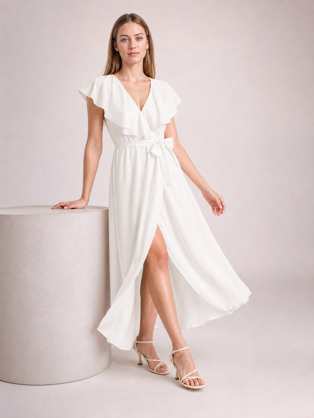 Model in a white ruffled surplice midi dress with flutter sleeves, a tied waist, flowing silhouette, and leg-baring front slit, posing with one hand on a pedestal.