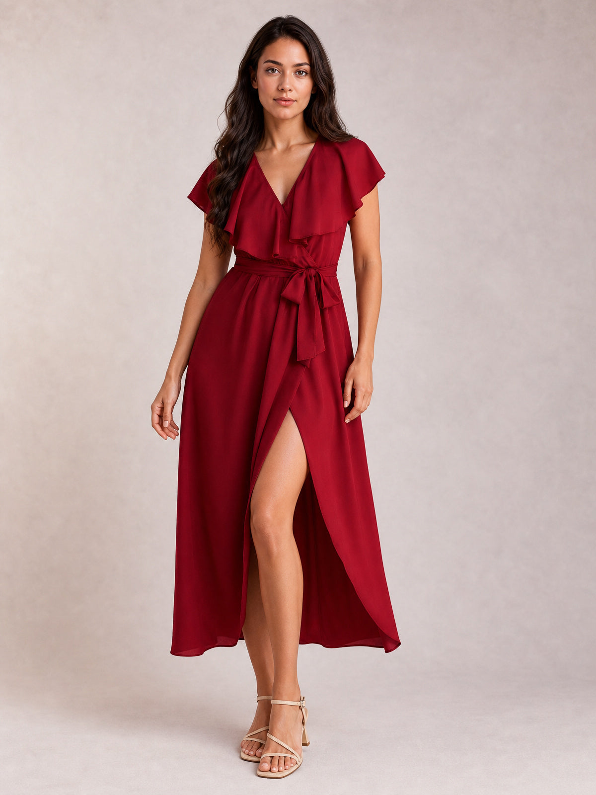 Model wears a burgundy ruffled surplice midi dress with flutter sleeves, a self-tie waist, flowing silhouette, and leg-baring front slit while standing.
