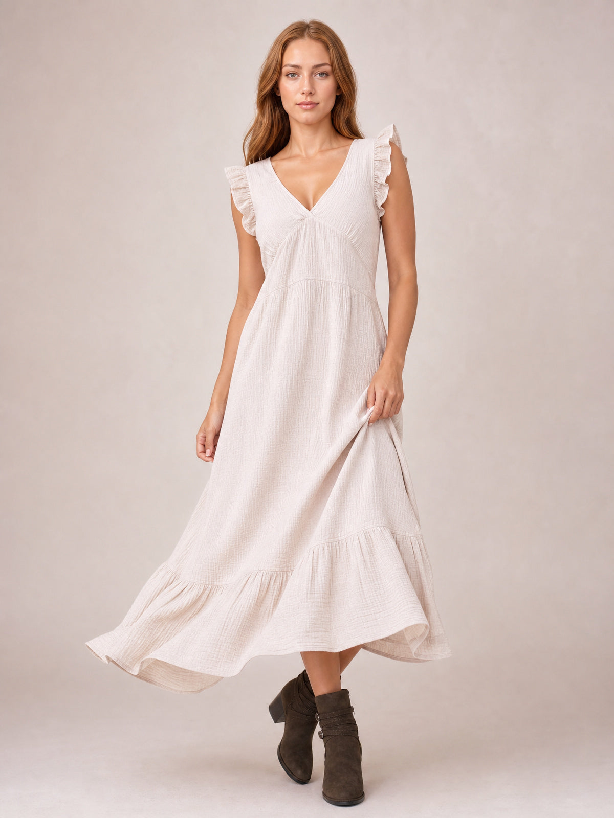 Model in a flowing cream ruffled V-neck maxi dress with cap sleeves, gathered waist, tiered hem, and side slit, lifting the skirt while posing in ankle boots.