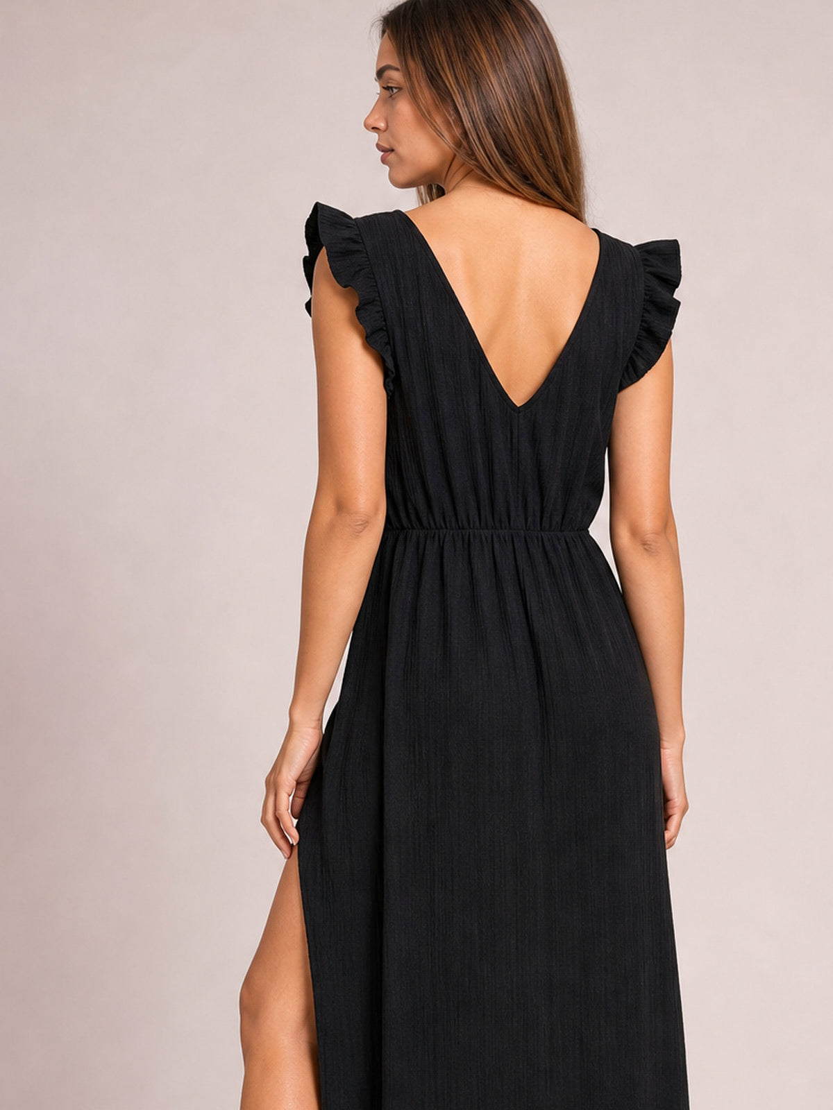 A model stands turned away in a black ruffled V-neck maxi dress with cap sleeves, gathered waist, flowing silhouette, deep V-back, and visible side slit.