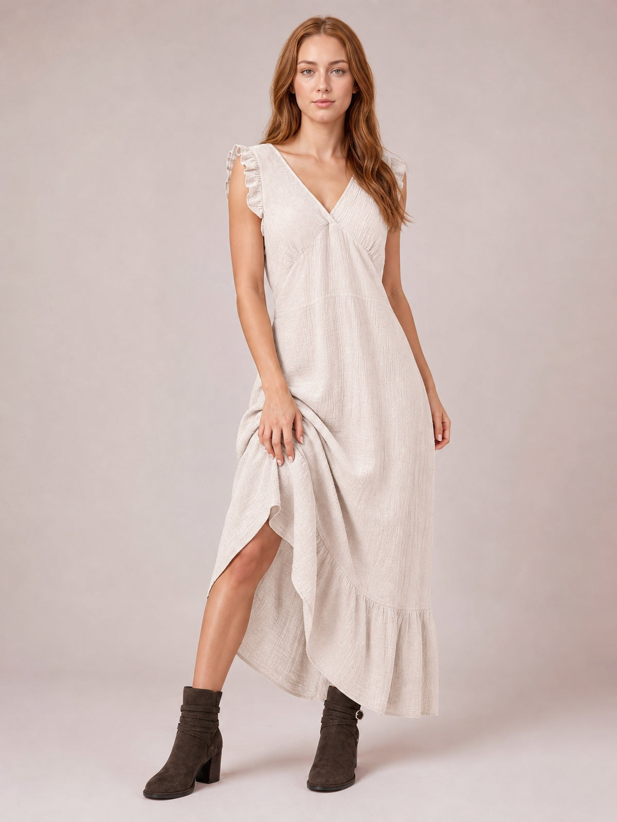 Model lifts an ivory ruffled V-neck maxi dress to reveal its side slit, gathered waist, flowing silhouette, cap sleeves, and tiered hem.