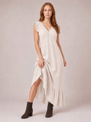 Model lifts an ivory ruffled V-neck maxi dress to reveal its side slit, gathered waist, flowing silhouette, cap sleeves, and tiered hem.
