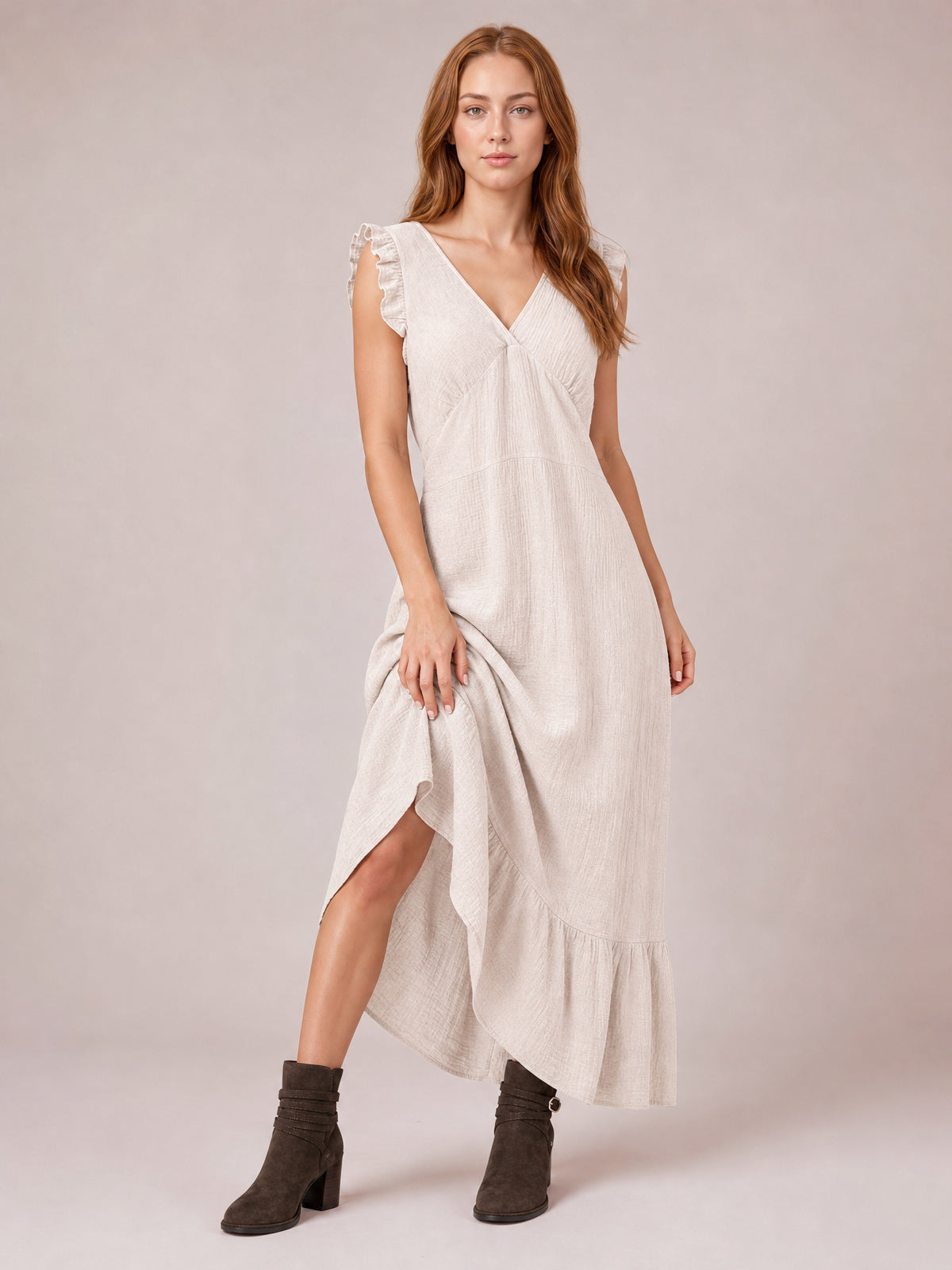 Model lifts an ivory ruffled V-neck maxi dress to reveal its side slit, gathered waist, flowing silhouette, cap sleeves, and tiered hem.
