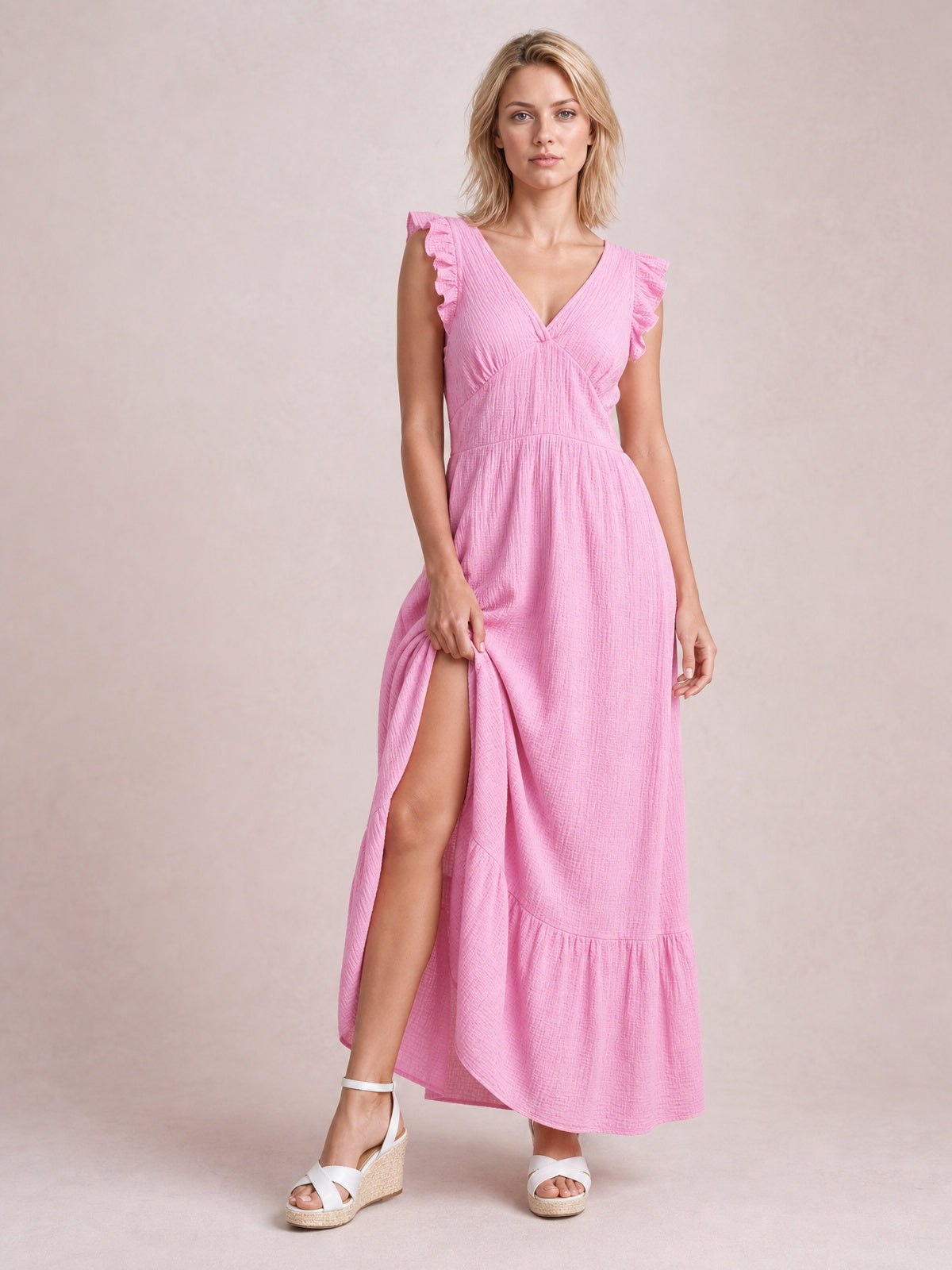 Model in a pink ruffled V-neck maxi dress with cap sleeves, a gathered waist, flowing tiered skirt, and visible side slit.