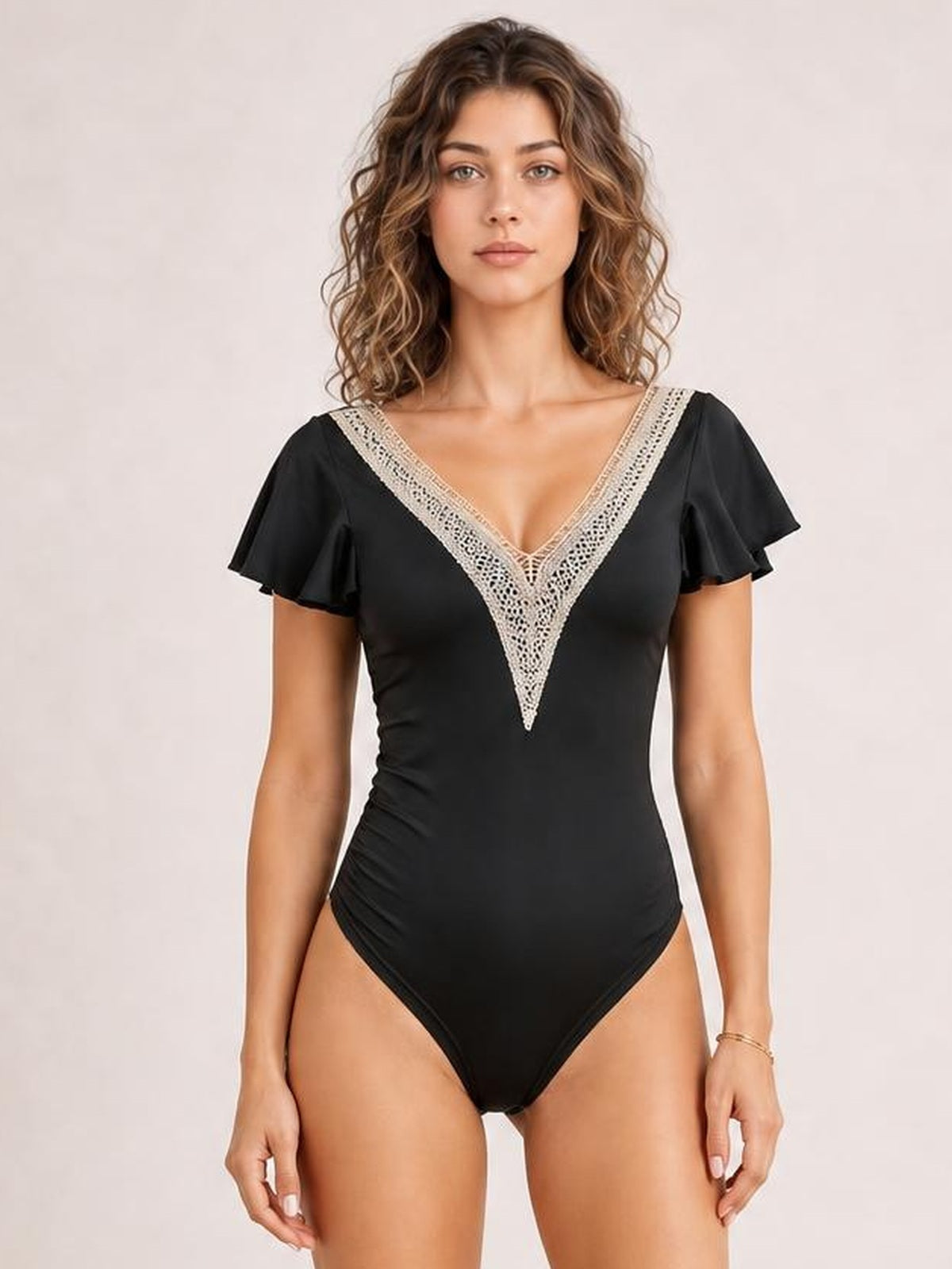 A model stands facing forward in a fitted black one-piece swimsuit with a deep V-neck, delicate lace trim, and ruffled cap sleeves.