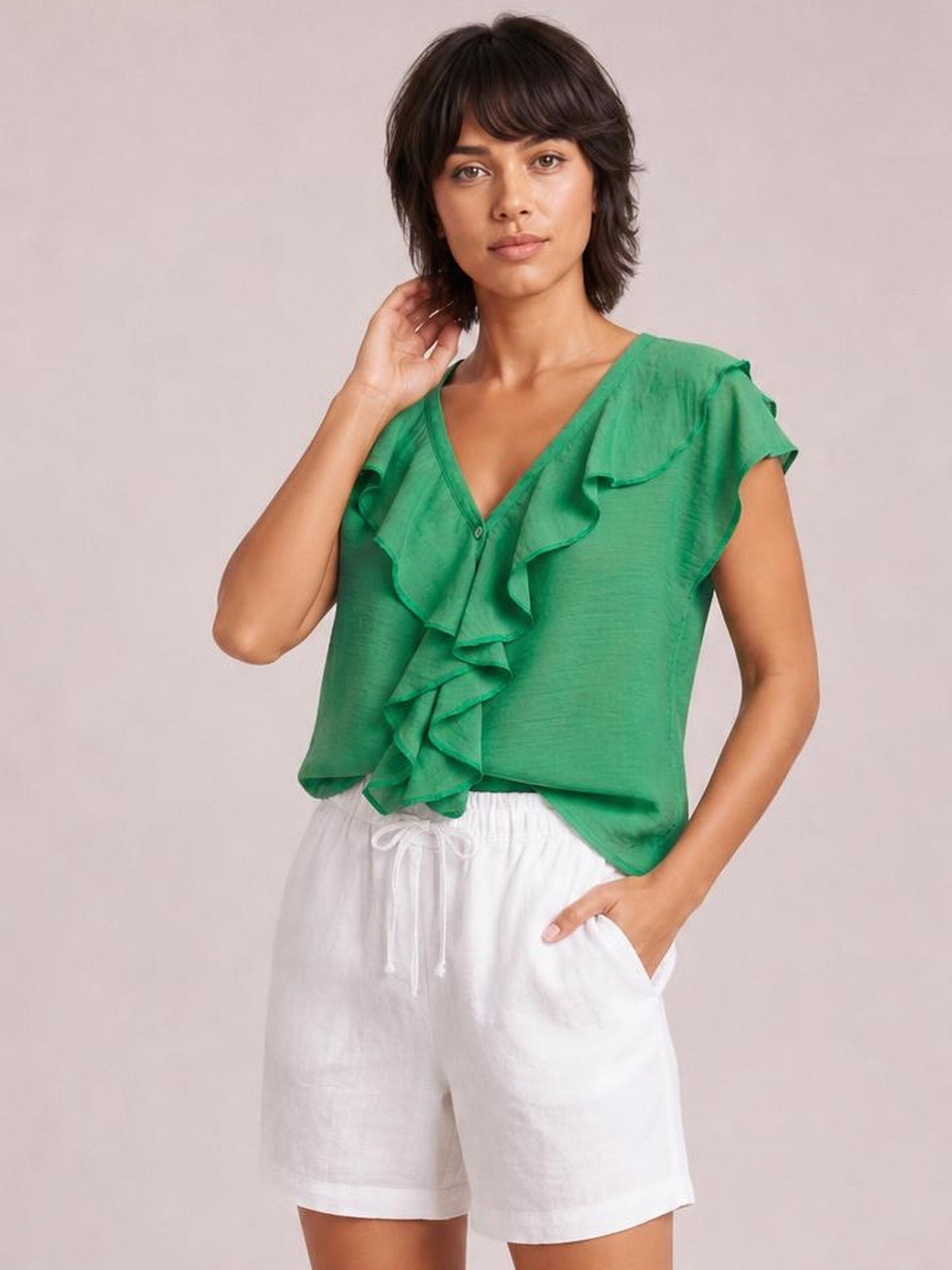 A model poses in a green ruffled V-neck short sleeve blouse with a relaxed silhouette, cascading front ruffles, and a visible button closure.