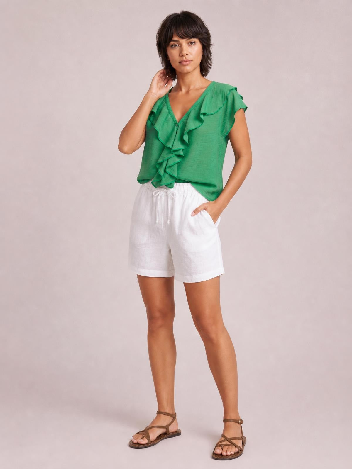 Model poses in a green ruffled V-neck short sleeve blouse with a relaxed silhouette, layered front ruffles, and a curved hem.