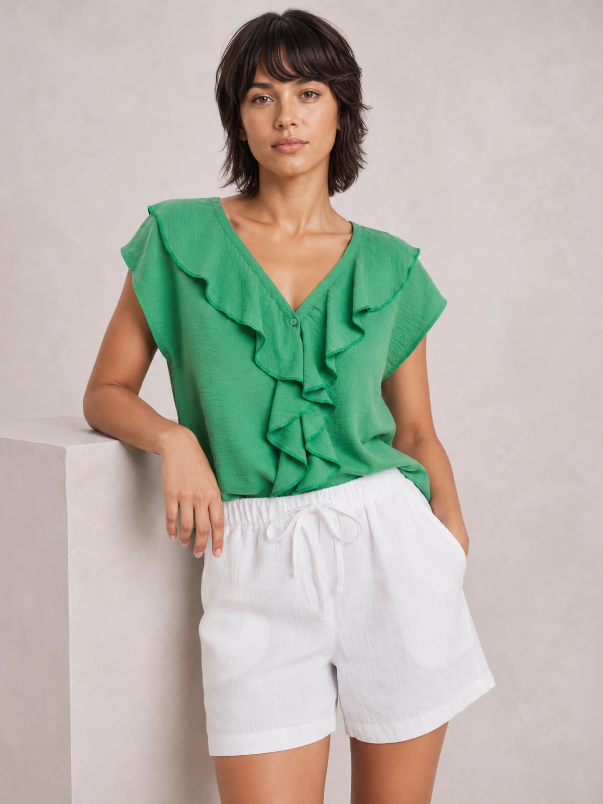 Model leans against a ledge wearing a green ruffled V-neck short sleeve blouse with a relaxed silhouette, cascading front ruffles, and button details.