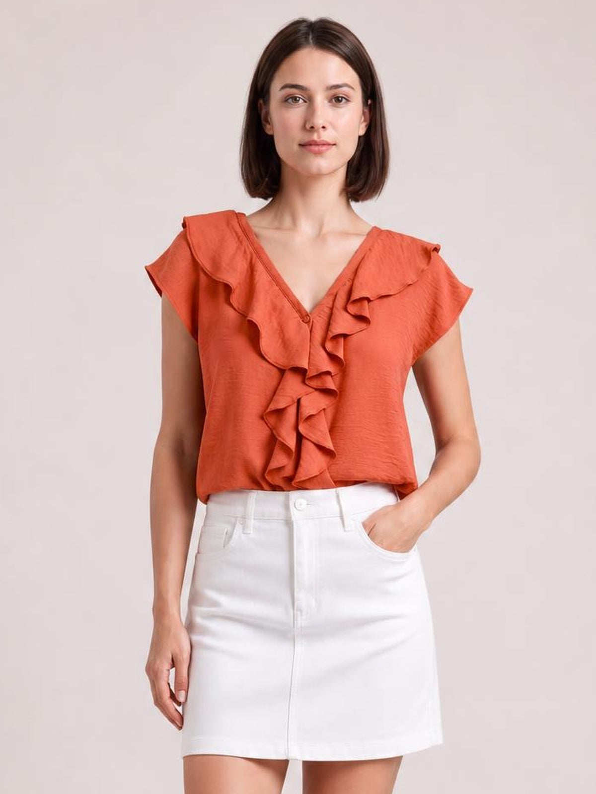 Model wearing a coral ruffled V-neck short sleeve blouse with a relaxed silhouette and cascading front ruffles, paired with a white skirt.