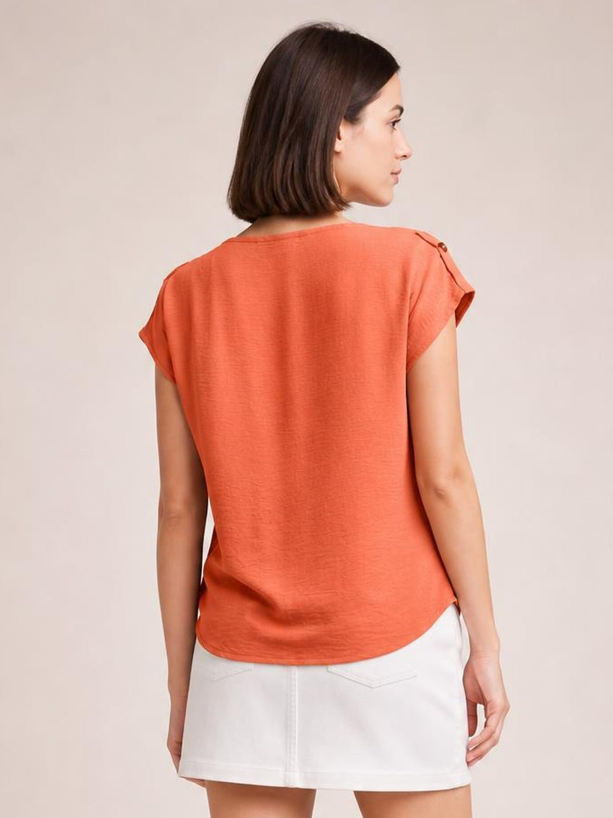 A model stands turned slightly away in a coral short-sleeve blouse with a relaxed silhouette, shoulder tab details, and a curved hem.