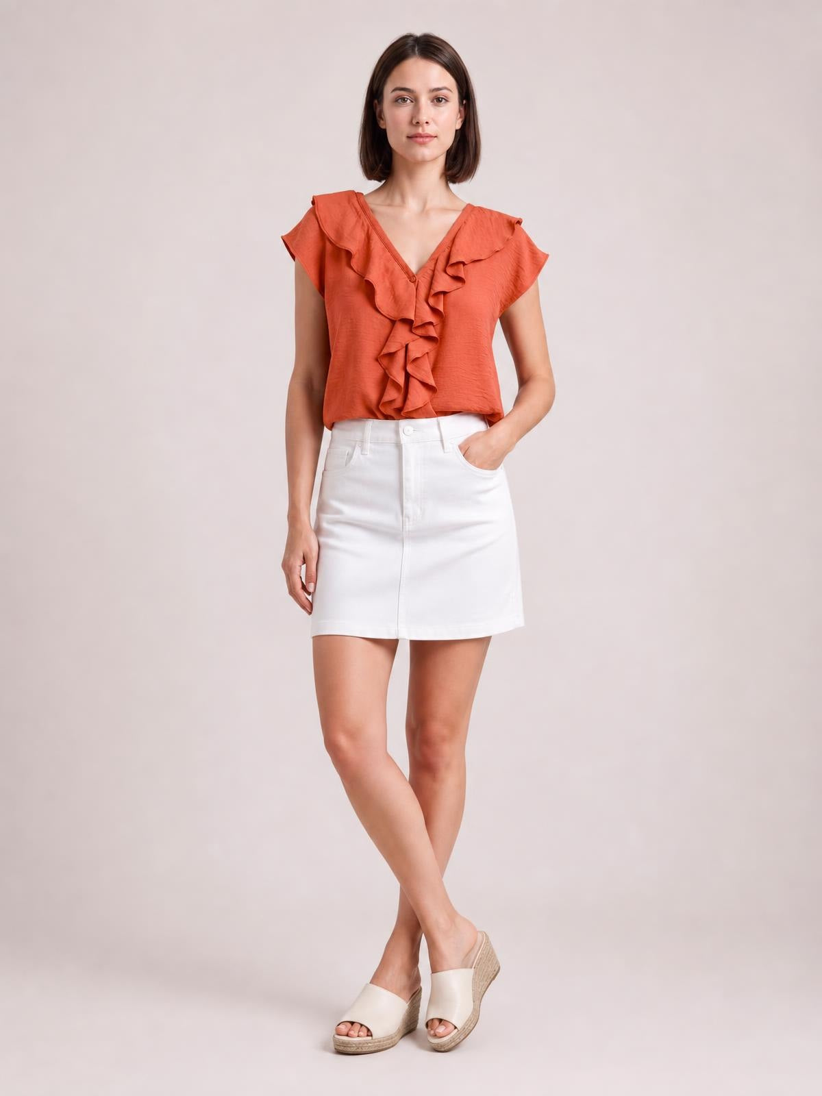 Model poses with one hand in her pocket wearing a coral ruffled V-neck short sleeve blouse with a relaxed silhouette and cascading front ruffles.