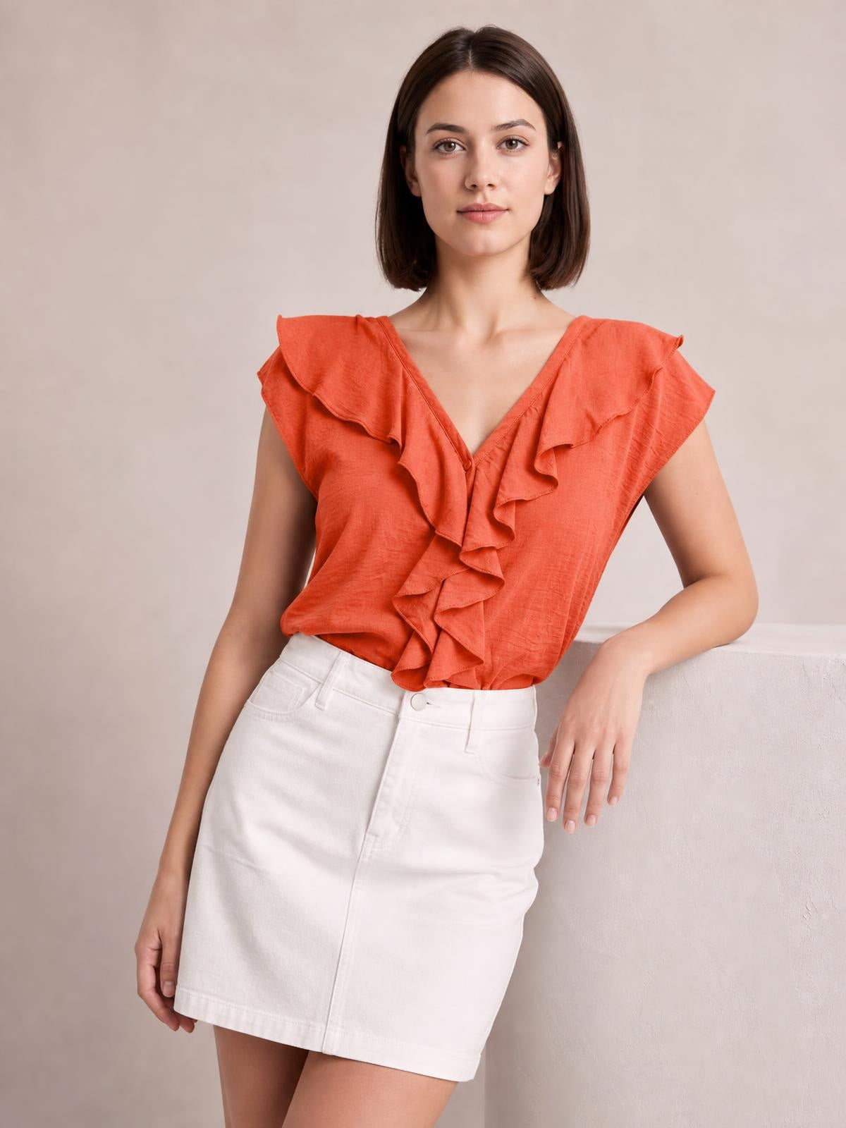 Model poses in an orange ruffled V-neck short sleeve blouse with a relaxed silhouette, layered ruffle detailing, and a softly draped hem.