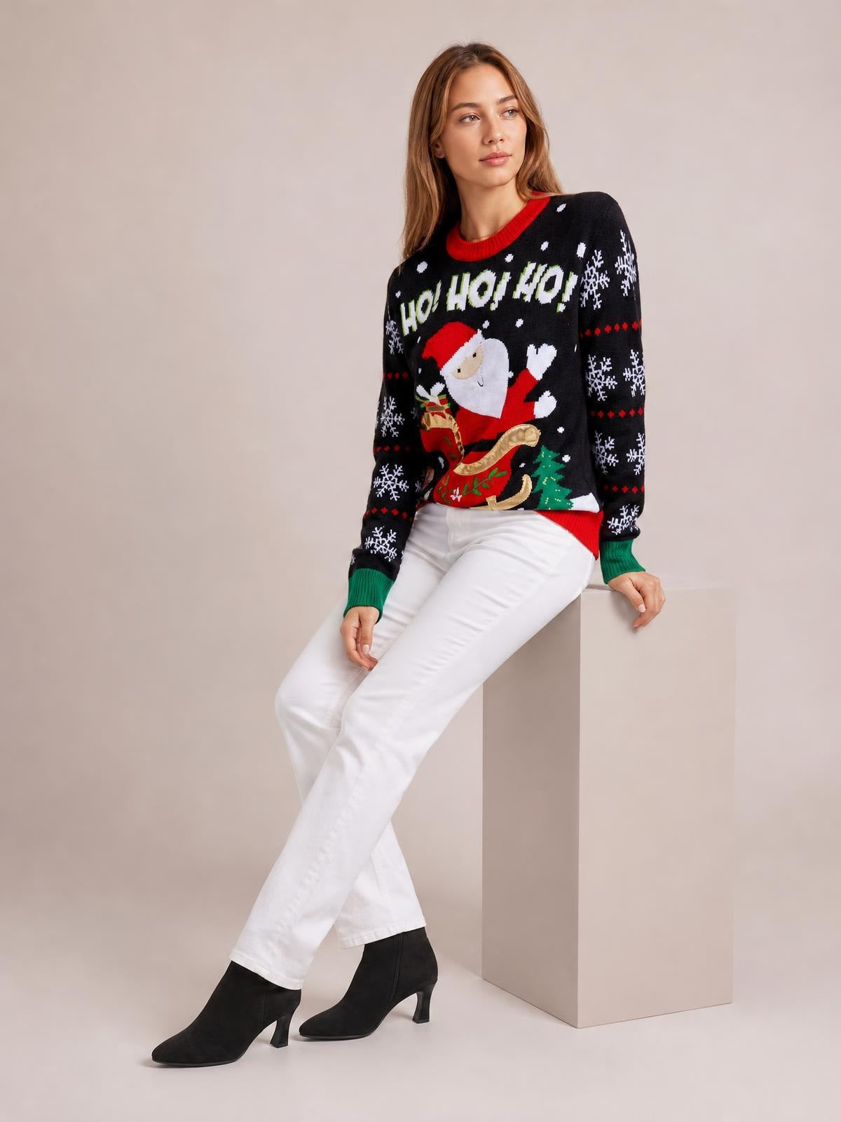 Model seated in a black Santa graphic round neck long sleeve sweater with red and green ribbed trim, relaxed silhouette, snowflake details, and festive front design.