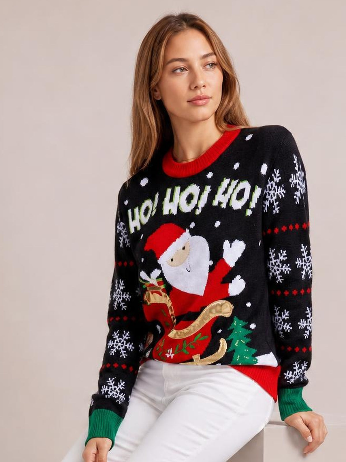 A seated woman wears a black Santa graphic round-neck long-sleeve sweater with snowflake motifs, red ribbed trim, green cuffs, and a relaxed silhouette.