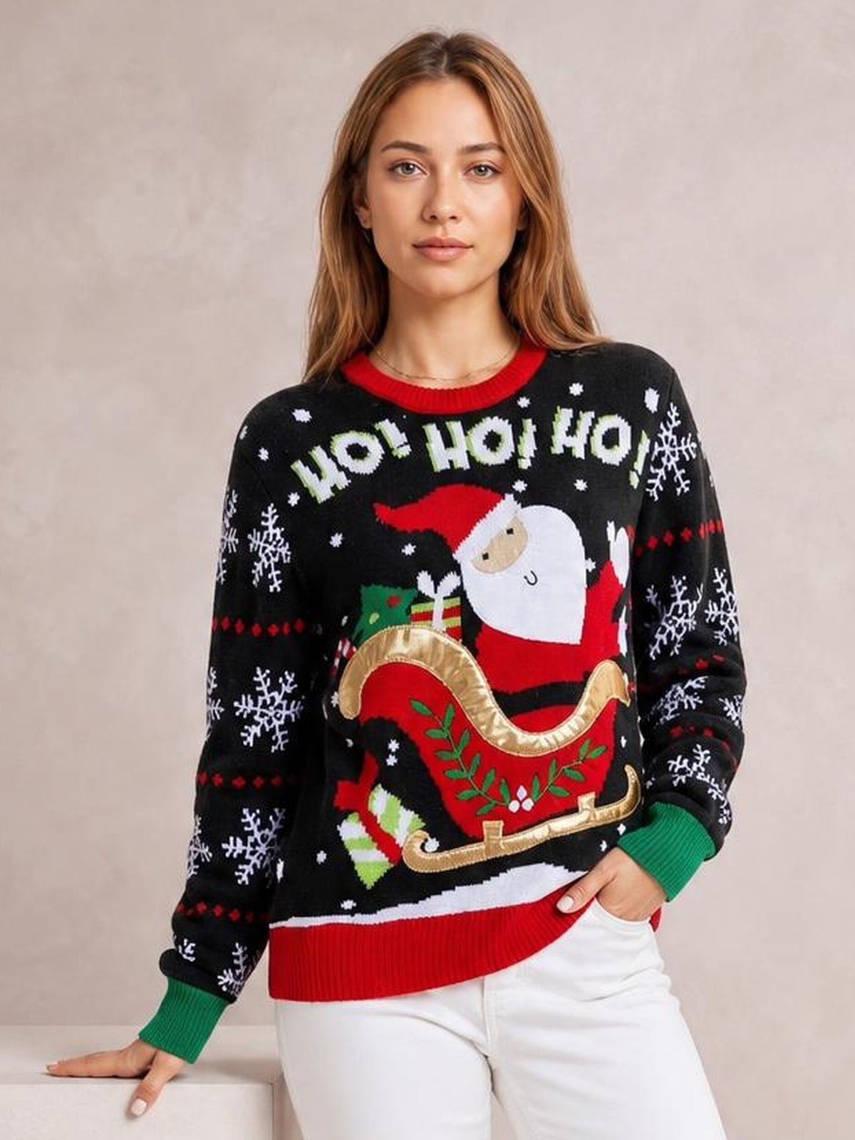 A model poses in a black Santa graphic round-neck long-sleeve sweater with ribbed red and green cuffs and hem, resting one hand on a ledge.