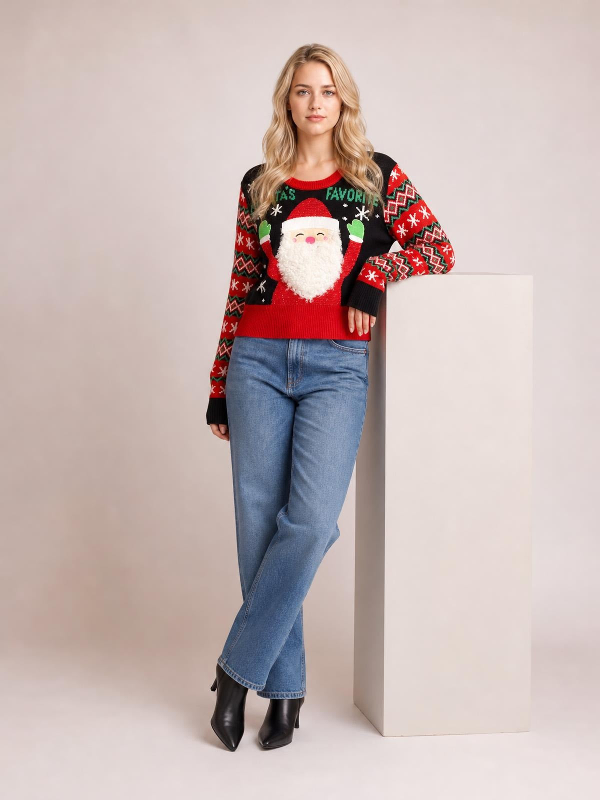 A model wears a black SANTA’S FAVORITE graphic long sleeve sweater with a Santa motif, red and green patterned sleeves, and ribbed trim, leaning with crossed legs.